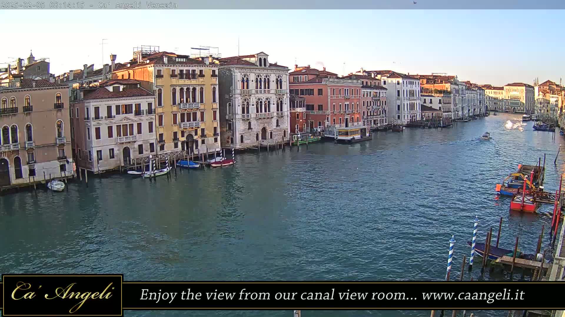A wide shot of a canal in Venice, Italy, shows several buildings lining the waterway, with boats moored along the sides and a few moving along the water under a clear, bright sky.