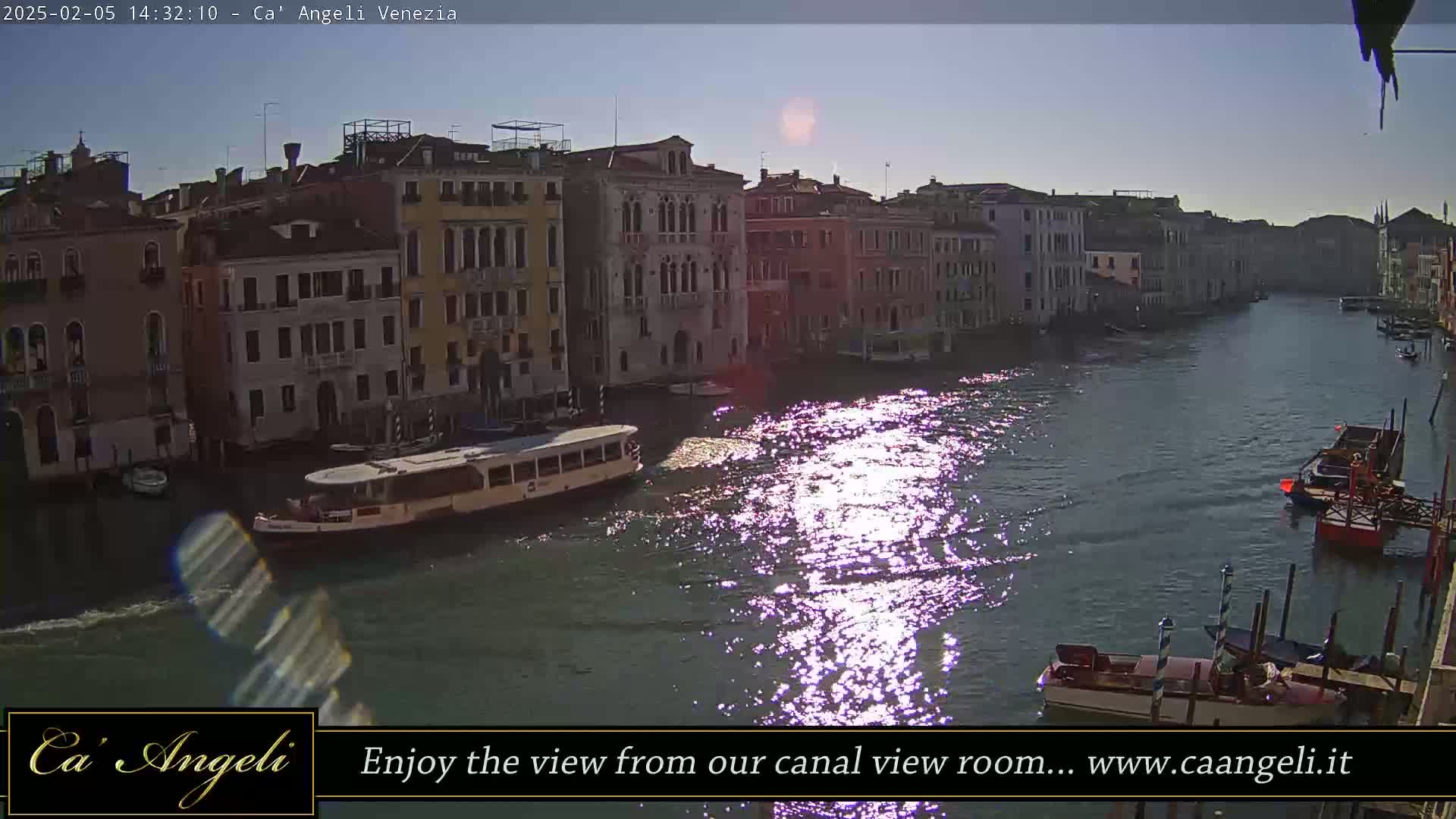 A sunny day in Venice shows a canal with several boats and buildings lining the waterway, sunlight reflecting on the water's surface.