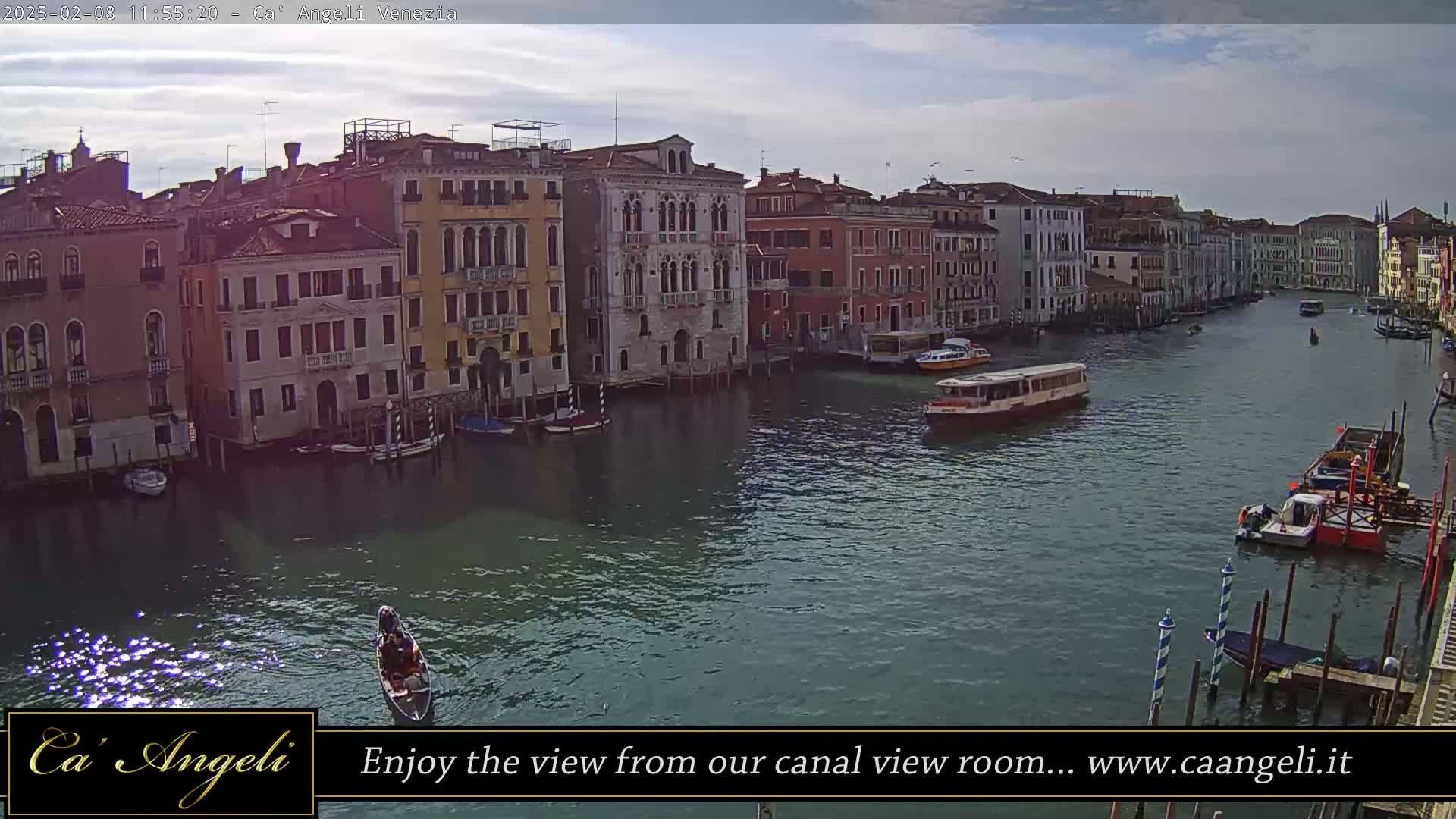 A gondola and several watercraft are on a wide canal lined with closely spaced, multi-story buildings under a partly cloudy sky.