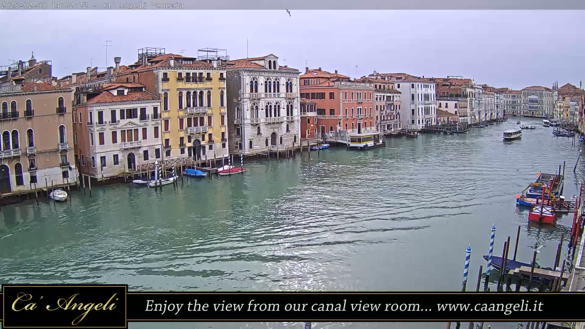 A wide shot shows a canal in Venice, Italy, with several multi-story buildings lining the waterway under an overcast sky; various boats are visible on the water.