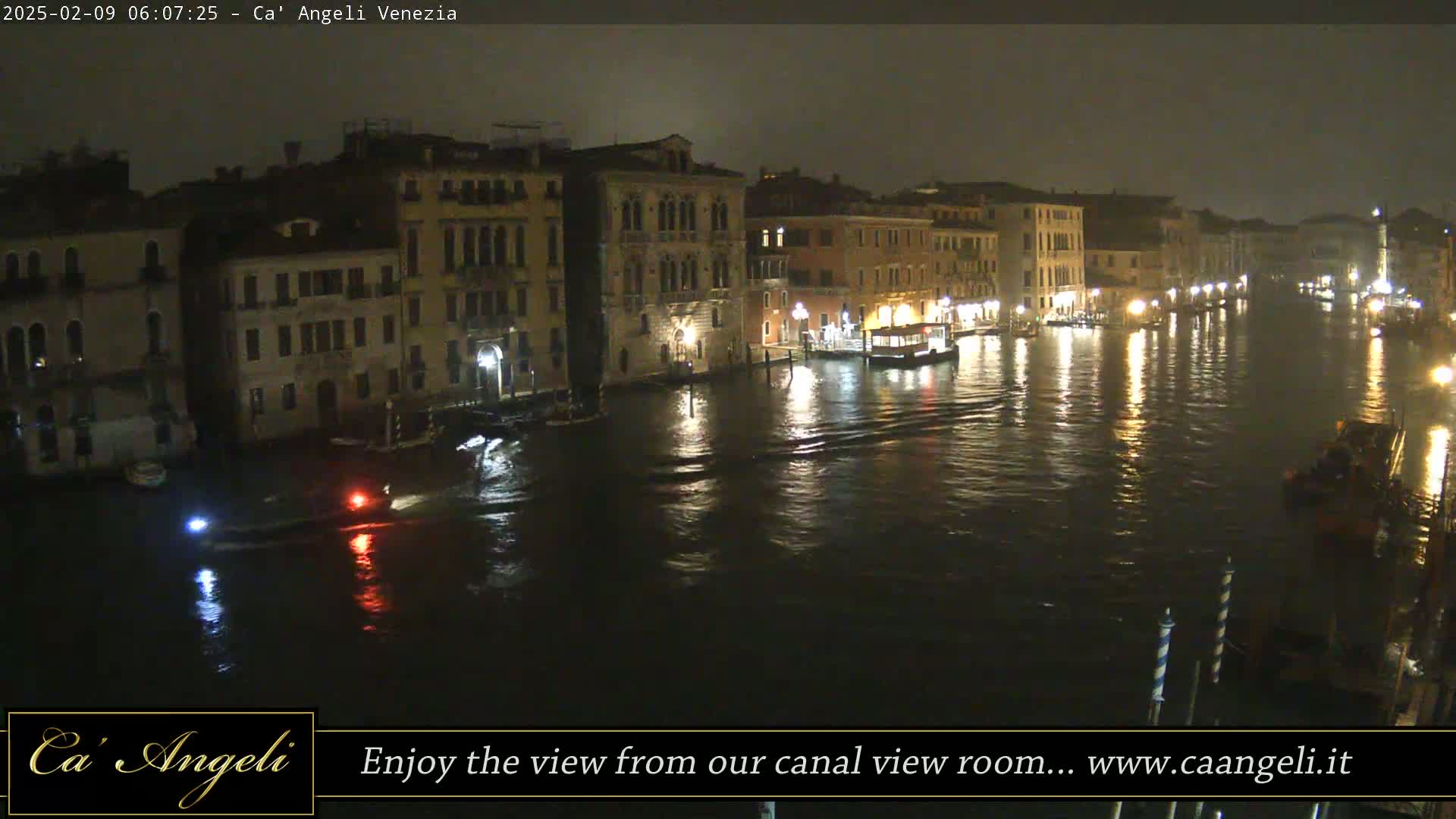At night, under a dark sky, a wide canal in Venice is lined with illuminated buildings, and small boats are visible on the water.