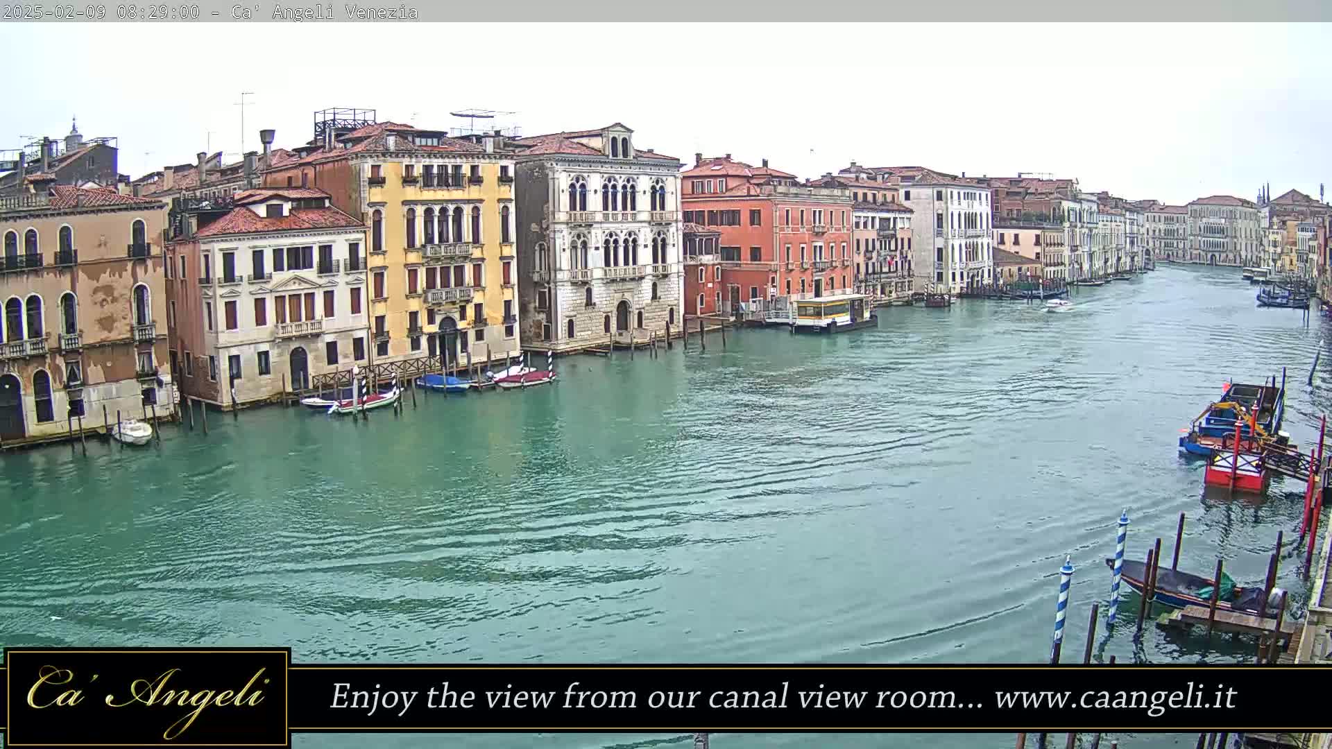 A wide canal in Venice, Italy, is shown on an overcast day, lined with colorful buildings and several boats.