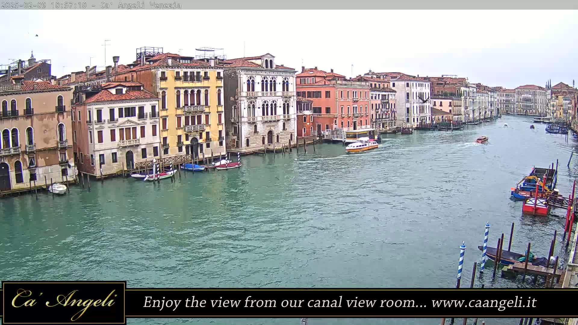 A wide shot shows a waterway with several boats and buildings along its edge under an overcast sky.