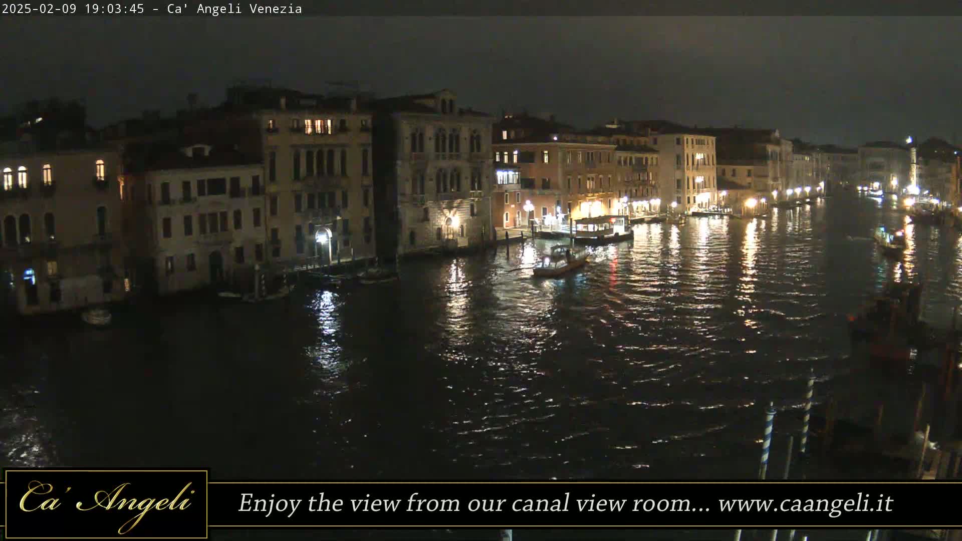A nighttime view of a canal in Venice, Italy, shows buildings lit along the waterway with boats on the water.