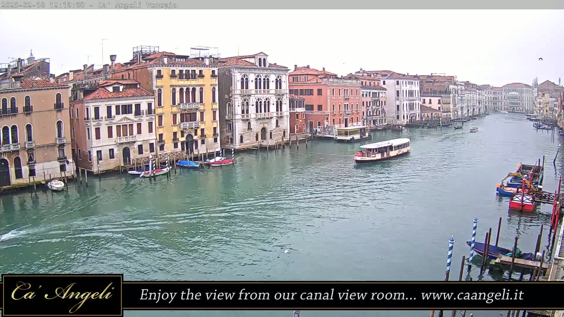 A wide shot of a canal in Venice, Italy, on an overcast day shows numerous buildings along the waterway with several boats and a water bus.