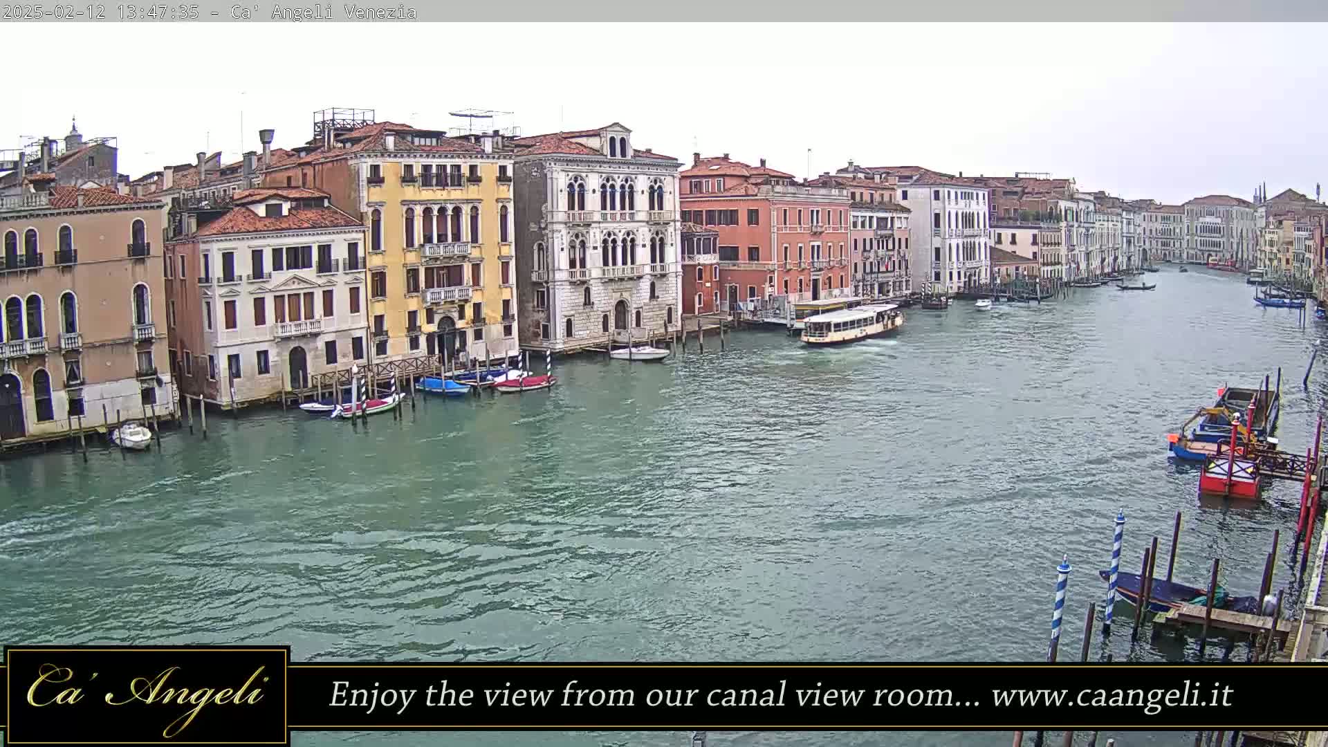 A wide shot of a canal in Venice, Italy, shows several buildings along the water's edge, numerous boats, and a mostly overcast sky.