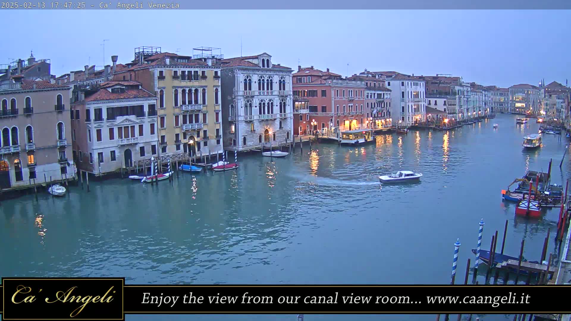 A twilight view of a canal in Venice, showing colorful buildings lining the waterway with boats moored along the sides and a few small boats traveling on the water.