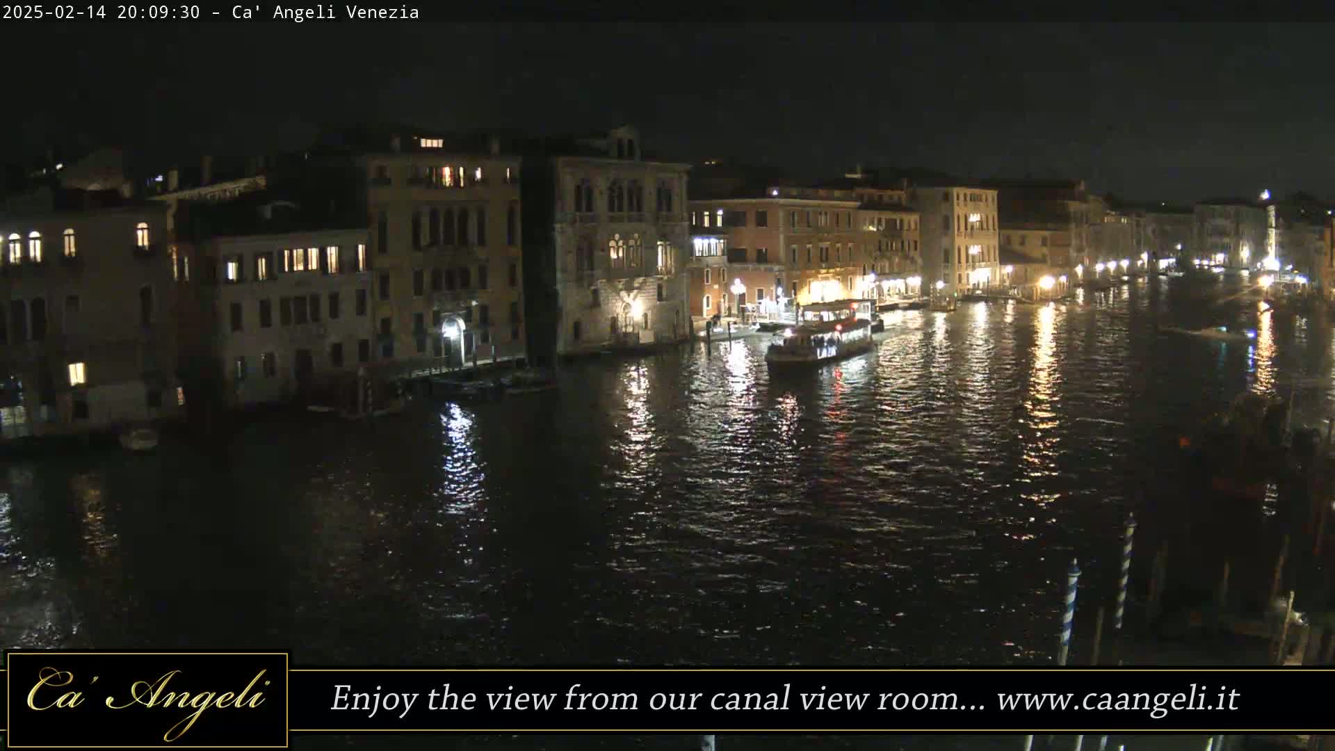 A nighttime view of a canal in Venice, Italy, shows illuminated buildings lining the waterway, with boats visible on the dark water.