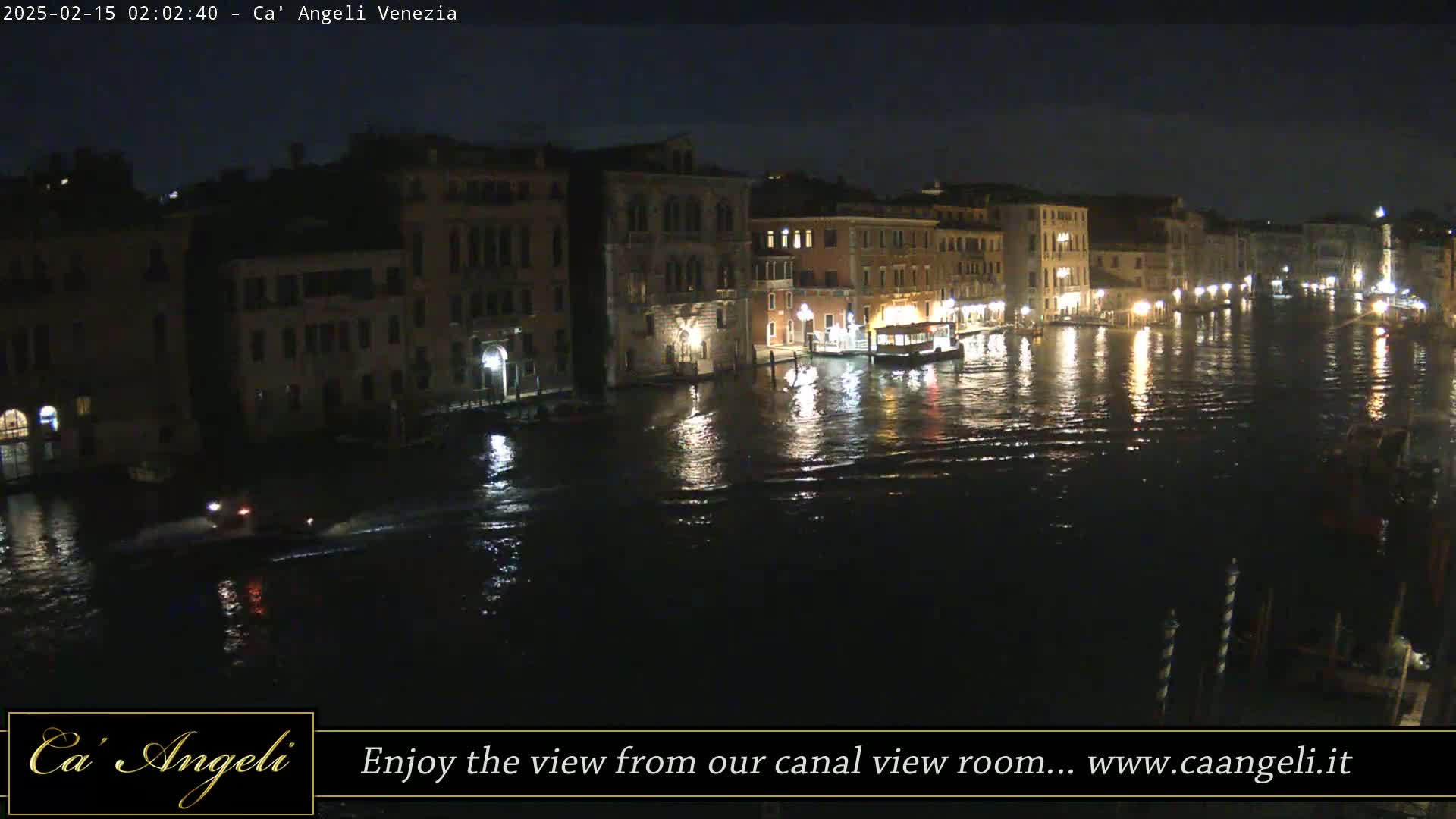 A nighttime view of a canal in Venice, Italy, with illuminated buildings lining the waterway and a small boat moving on the dark water.