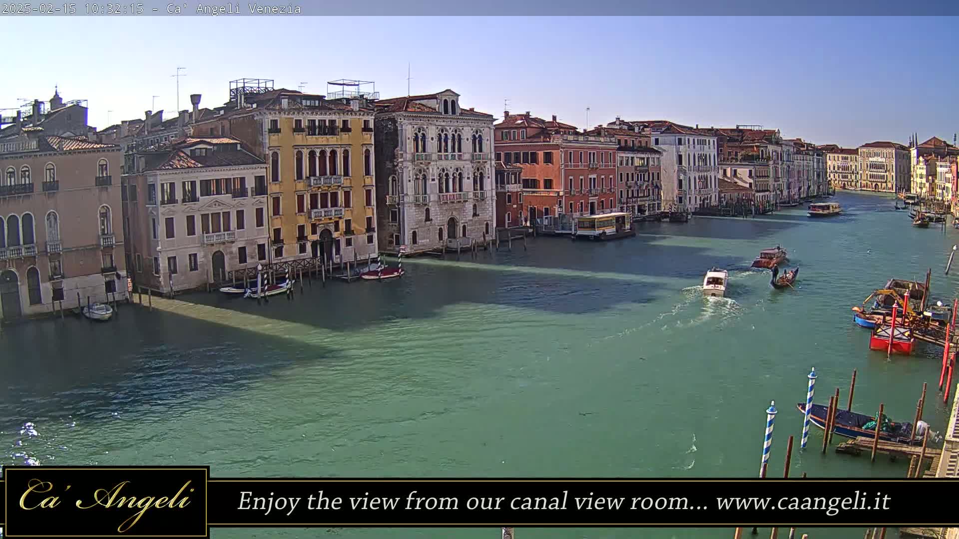 A sunny day in Venice shows a wide canal with colorful buildings lining the waterfront, several boats are visible on the water.