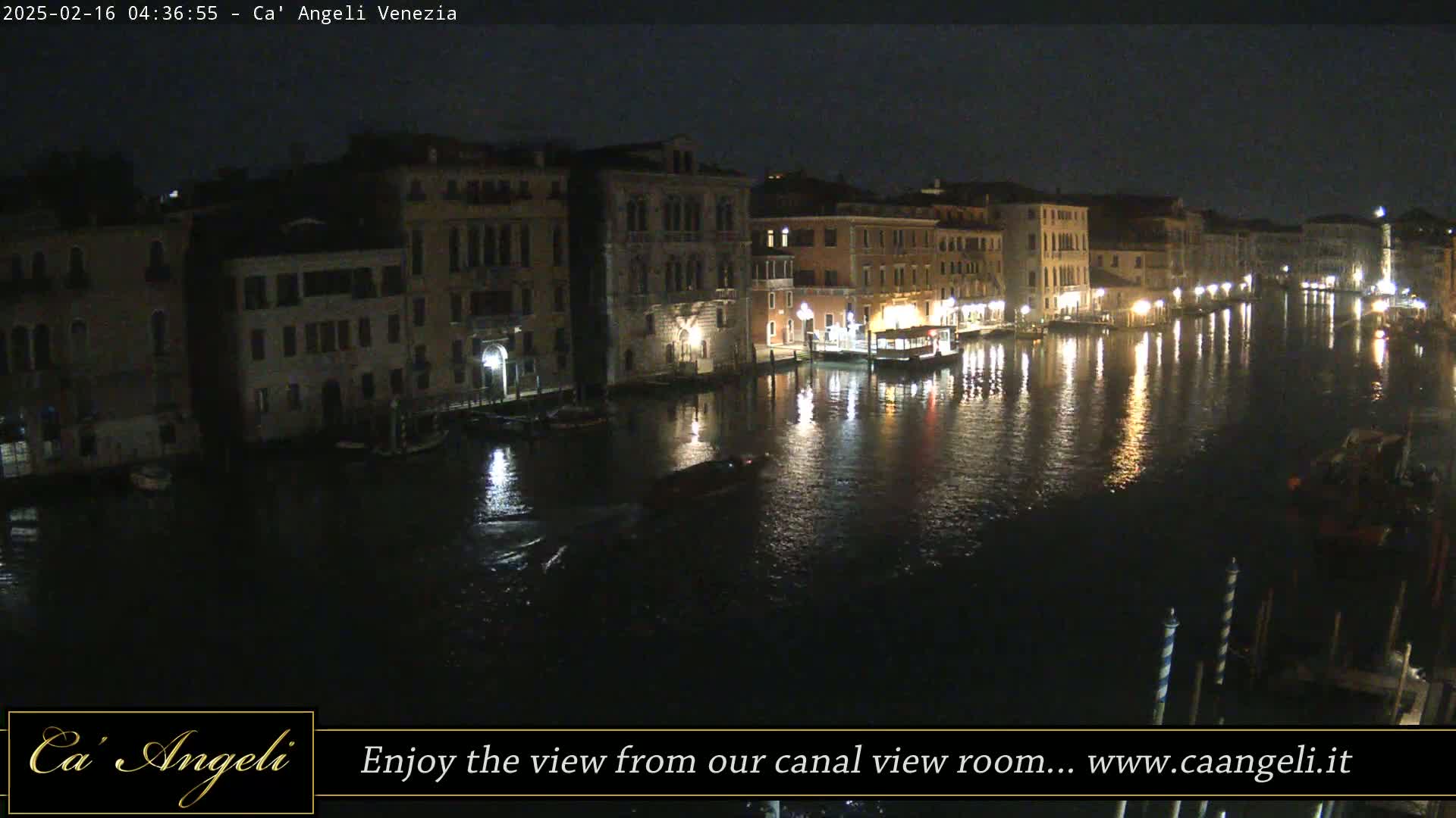 A nighttime view of a canal in Venice, Italy, showing illuminated buildings lining the waterway and boats on the water.