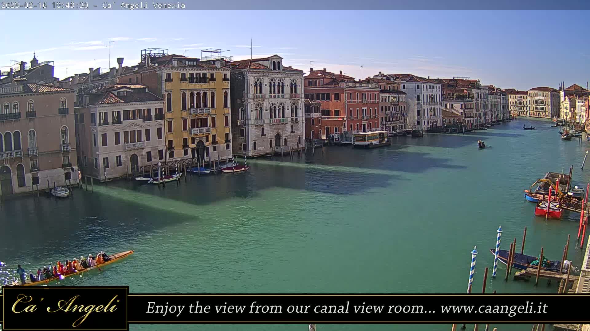 A sunny day in Venice shows a canal lined with colorful buildings, several boats, and a long boat with many people rowing.