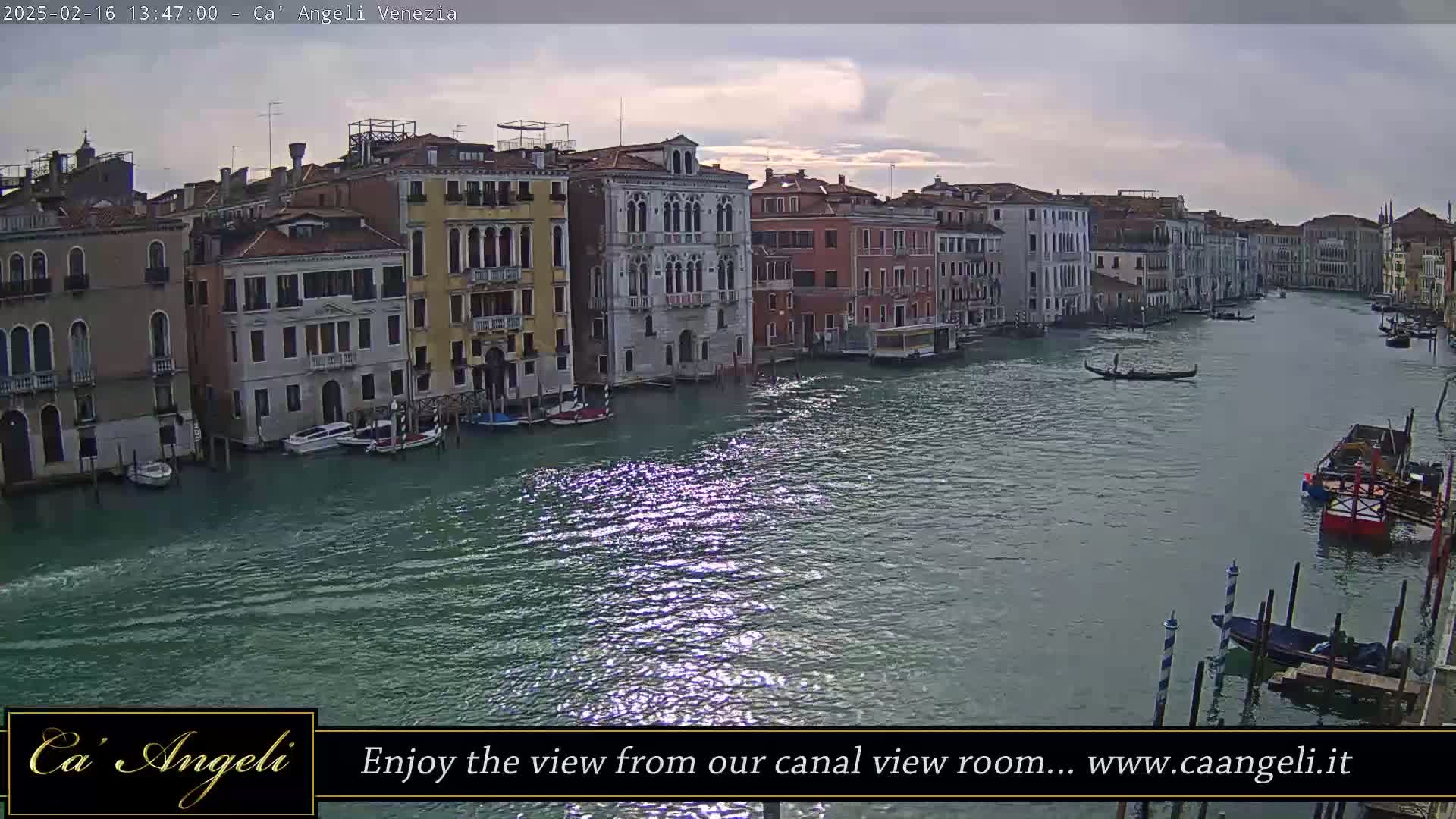 A wide shot of a canal in Venice, under a cloudy sky, shows numerous buildings lining the waterway with several boats moored along the banks and a gondola on the water.