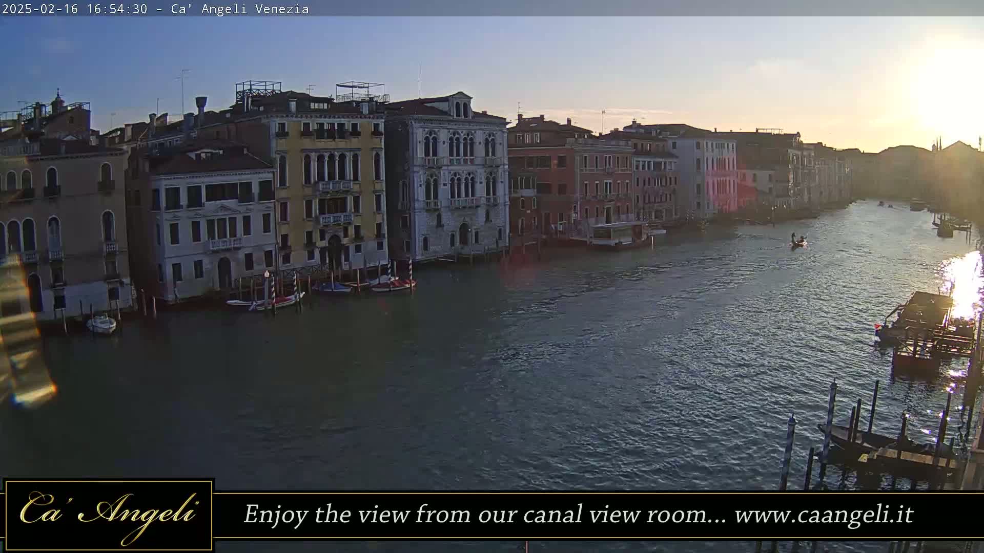 A wide shot of a Venetian canal at sunset, showing colorful buildings lining the waterway with gondolas and boats floating on the water.