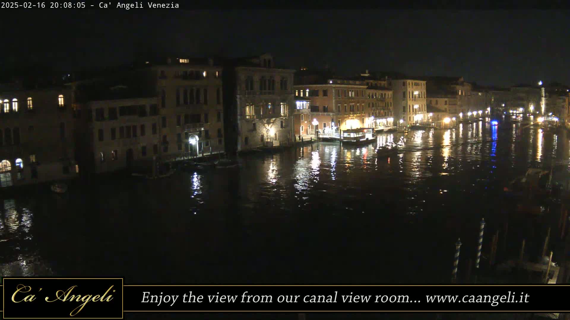 A nighttime view of a canal in Venice, Italy, shows illuminated buildings lining the waterway, with boats and reflections on the dark water.