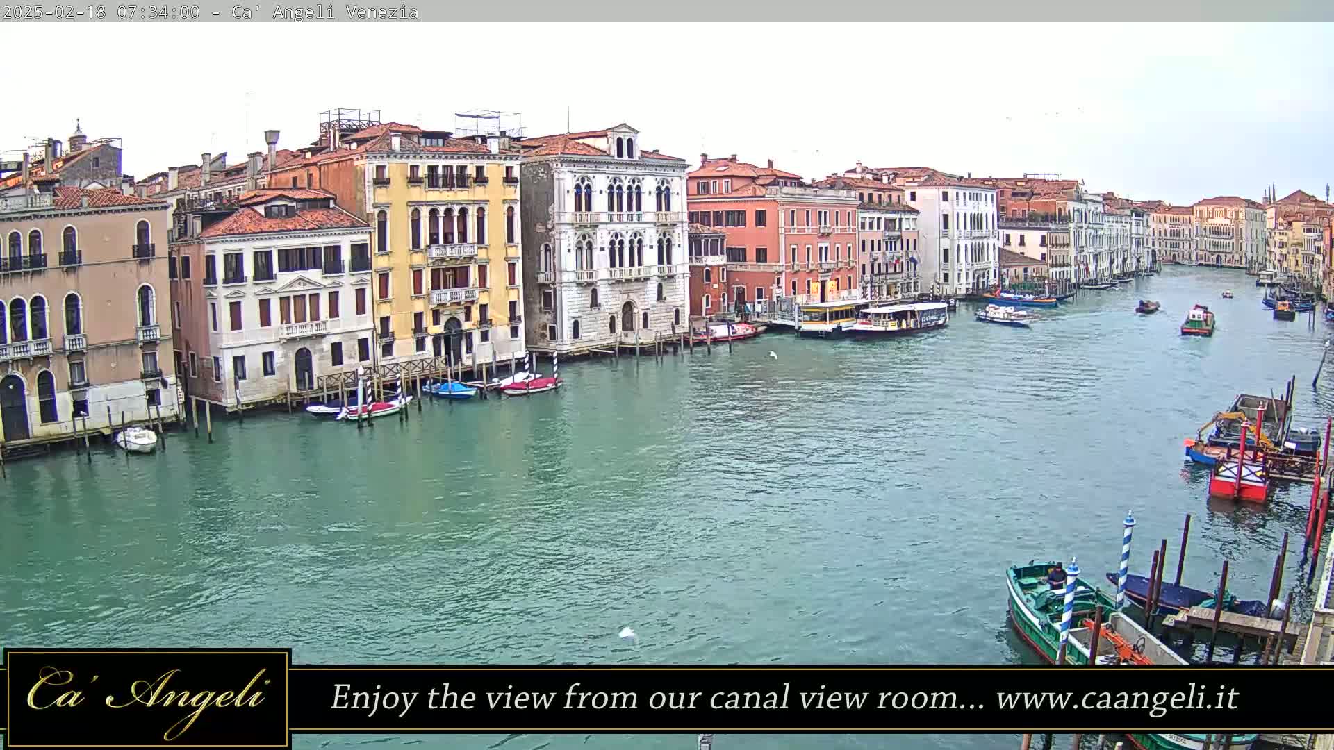 A wide shot of a waterway in Venice, Italy, shows several multi-story buildings lining the canal under an overcast sky, with various boats moored along the shore and others moving along the water.