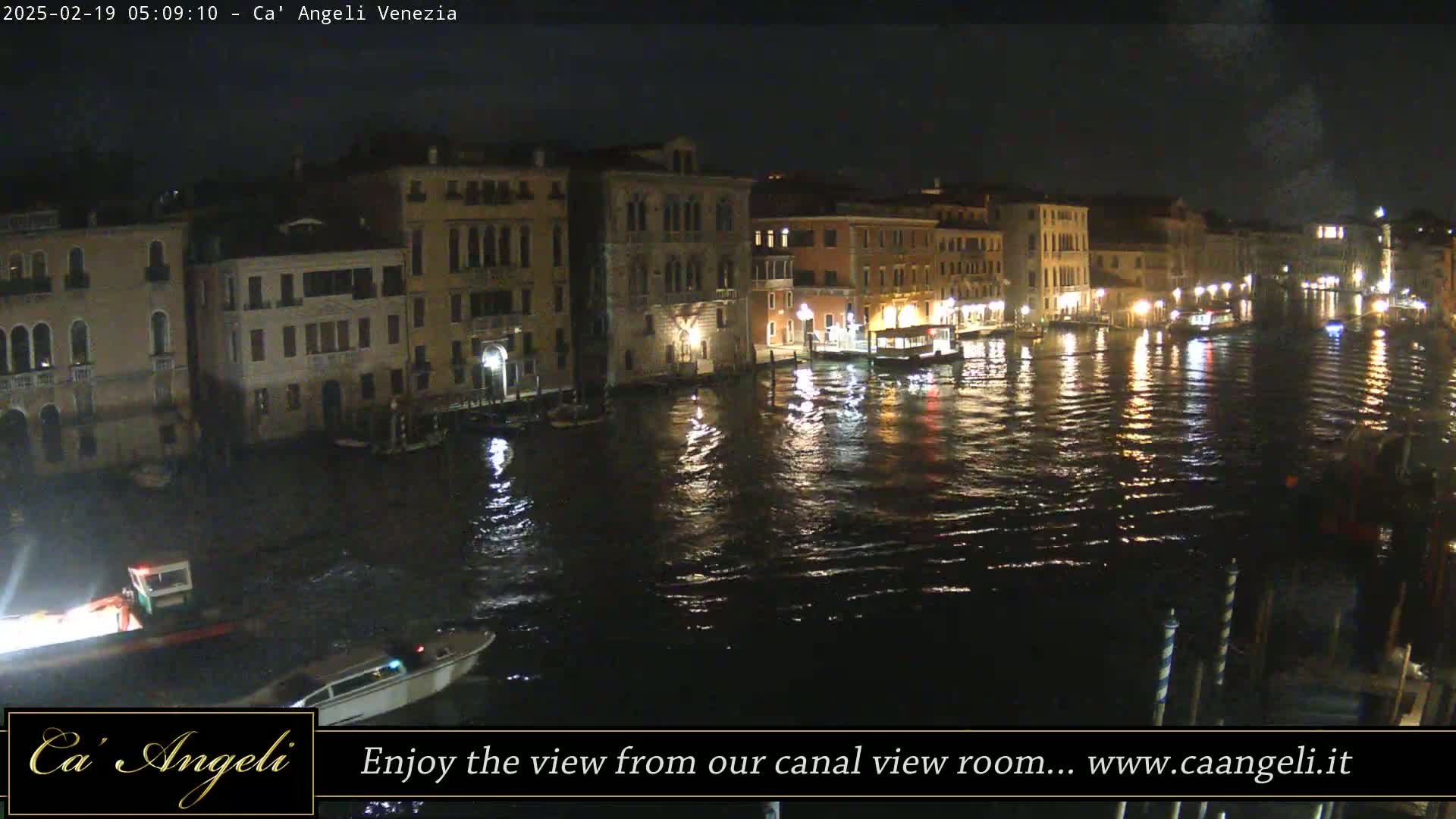 A nighttime view of a canal in Venice, showing illuminated buildings lining the waterway and boats on the water.