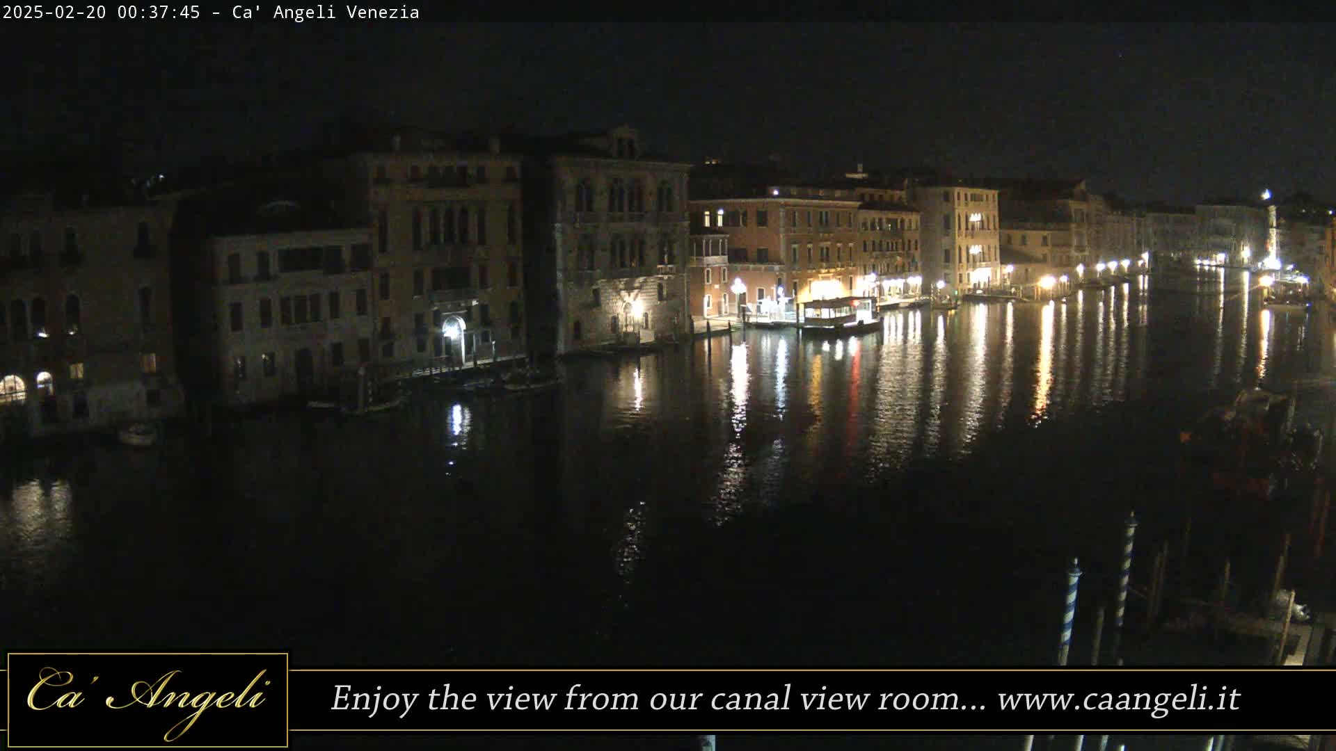 A nighttime view of a canal in Venice, Italy, shows illuminated buildings lining the waterway, with their lights reflecting on the dark water.