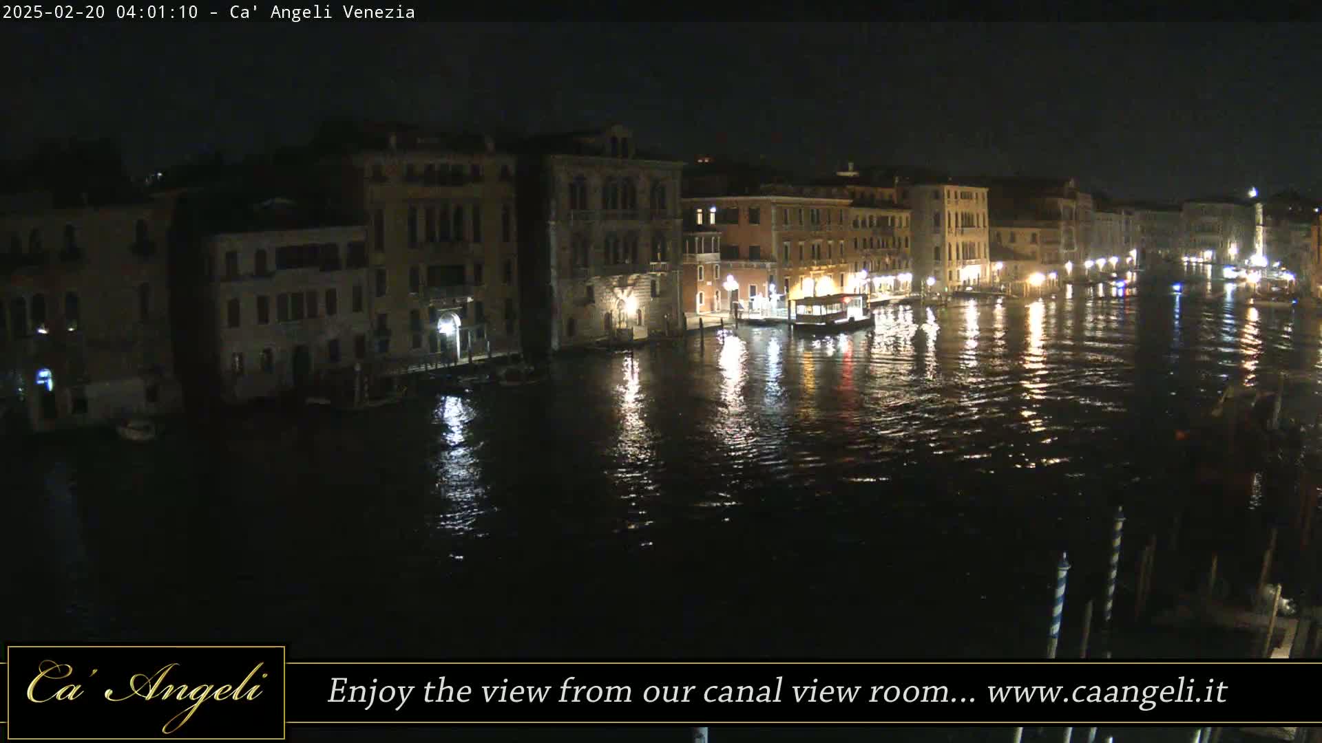 A nighttime view of a canal in Venice, showing illuminated buildings along the water's edge and several boats.