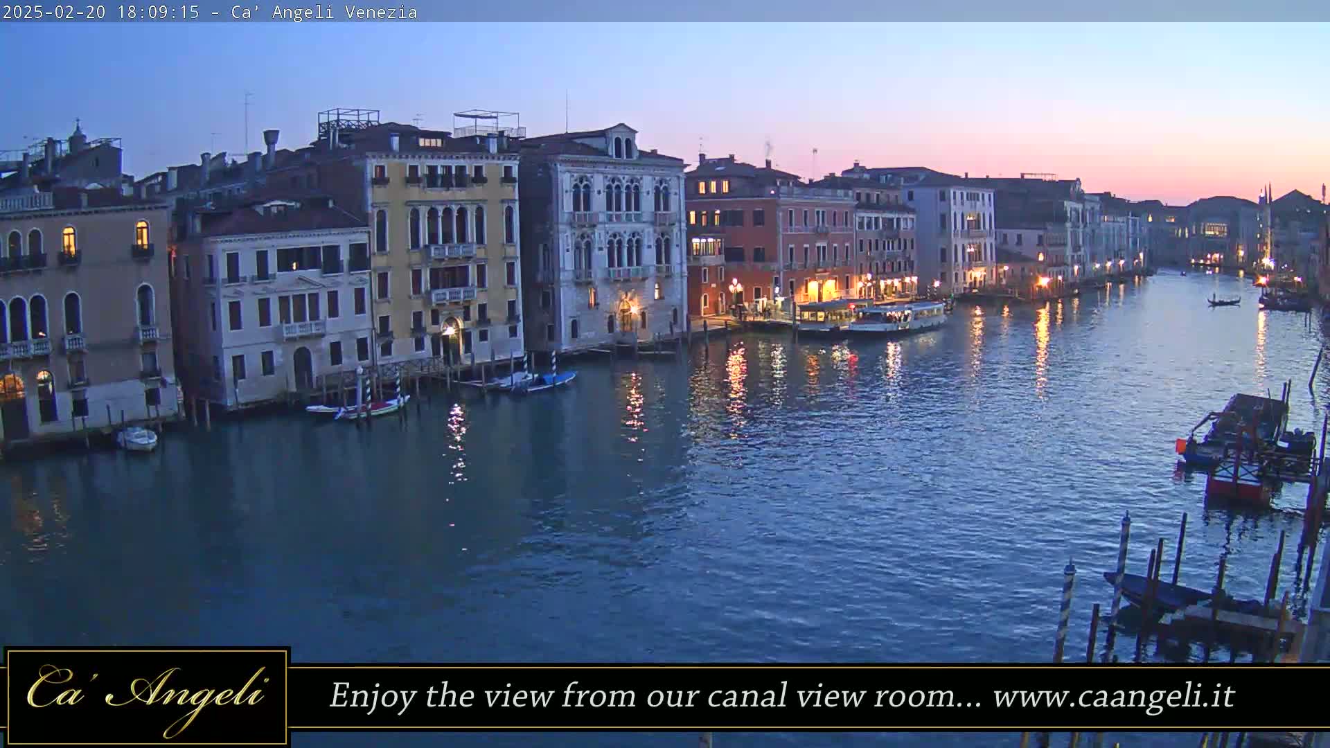 A wide canal in Venice at dusk, lined with illuminated buildings and boats, under a twilight sky.