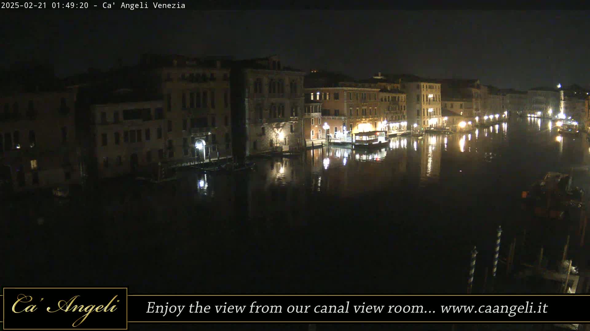 A nighttime view of a canal in Venice, showing illuminated buildings lining the waterway and their reflections in the dark water.