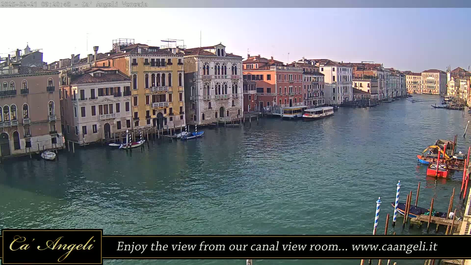A wide shot of a canal in Venice, Italy, on a clear day, shows numerous buildings lining the waterway with boats moored along the banks.