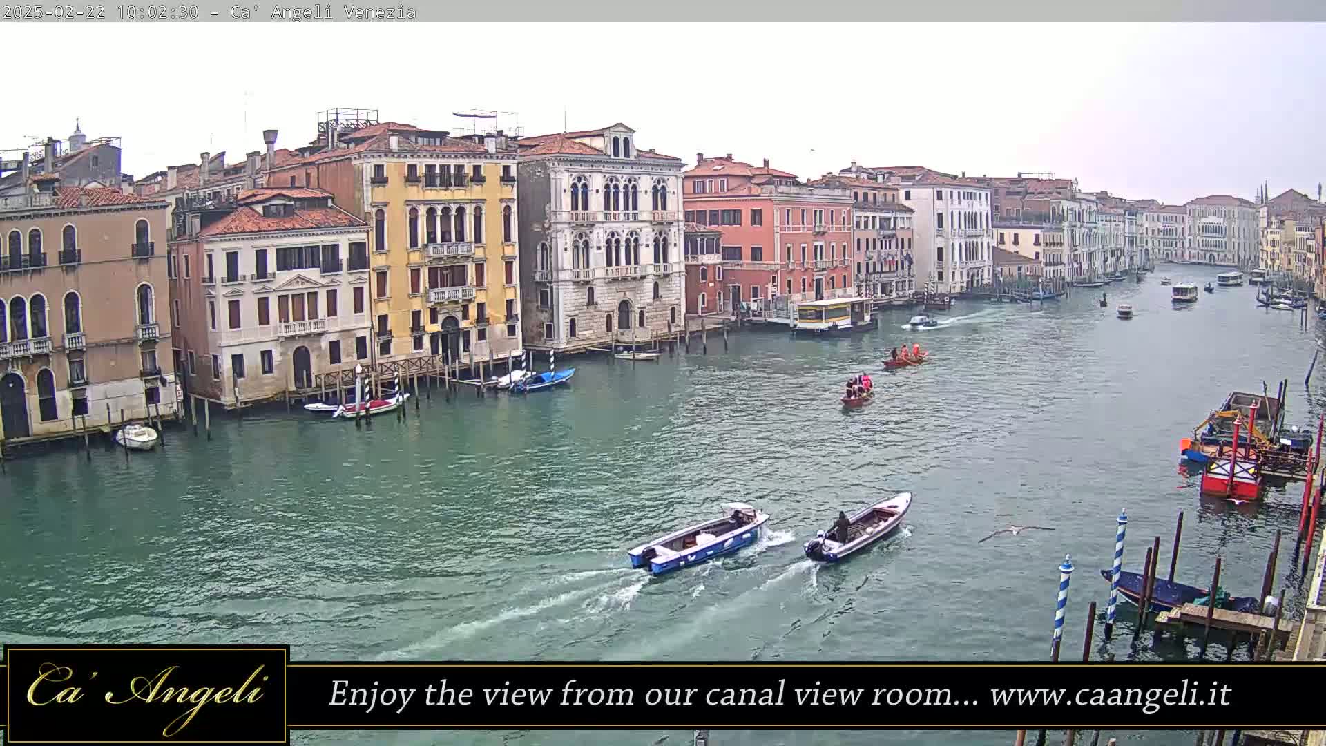 A wide shot of a canal in Venice on an overcast day, showing numerous buildings lining the waterway and several boats traveling along it.