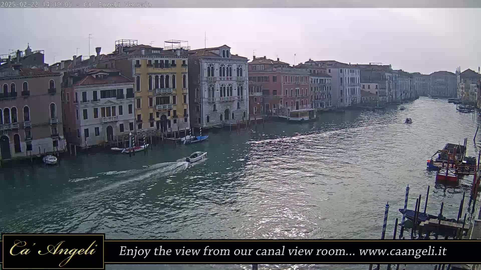 A wide shot shows a calm canal in Venice, Italy, under an overcast sky, with several buildings lining the waterway and several boats traveling along it.