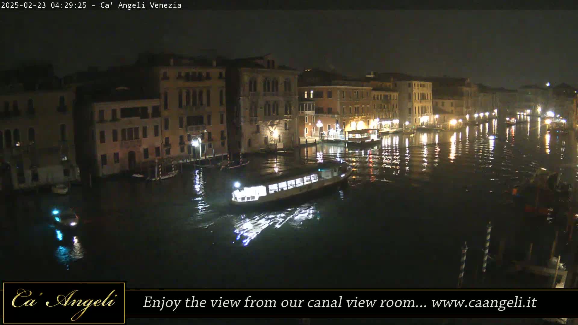 At night, a canal in Venice is lit by building lights reflected on the water, with a boat passing by.
