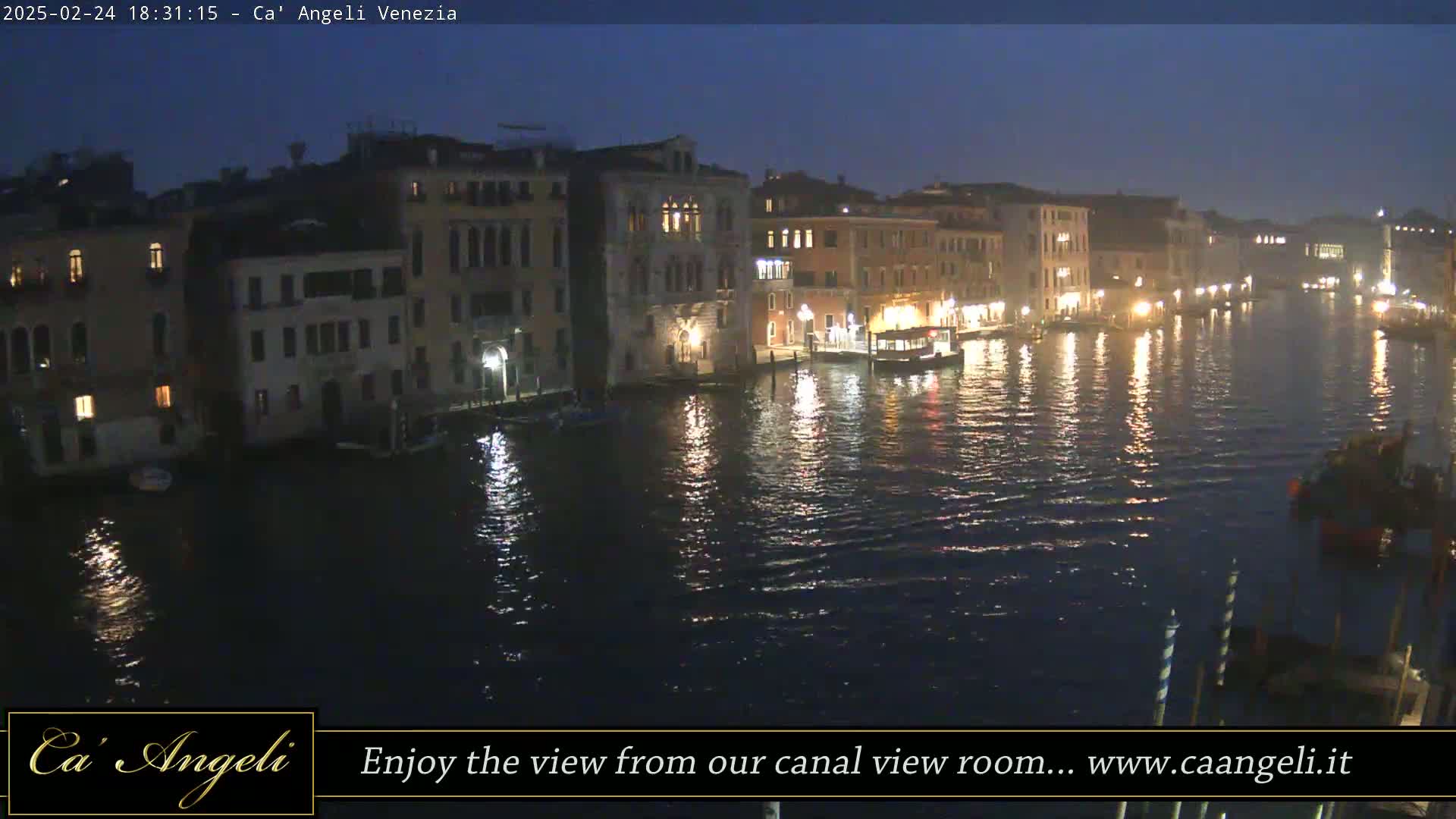 A nighttime view of a canal in Venice, Italy, shows illuminated buildings lining the waterway, with boats and calm water reflecting the lights.