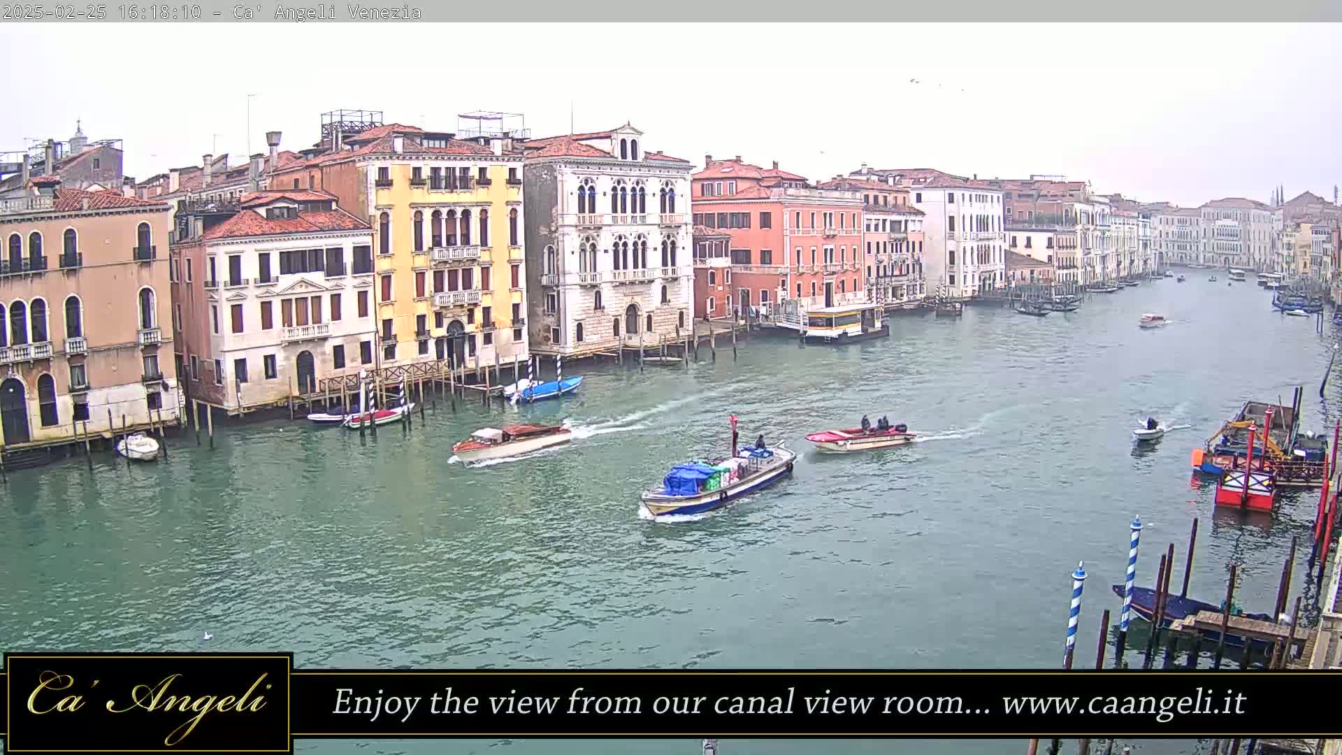 Several boats travel along a canal in Venice on an overcast day, lined with closely-packed buildings.