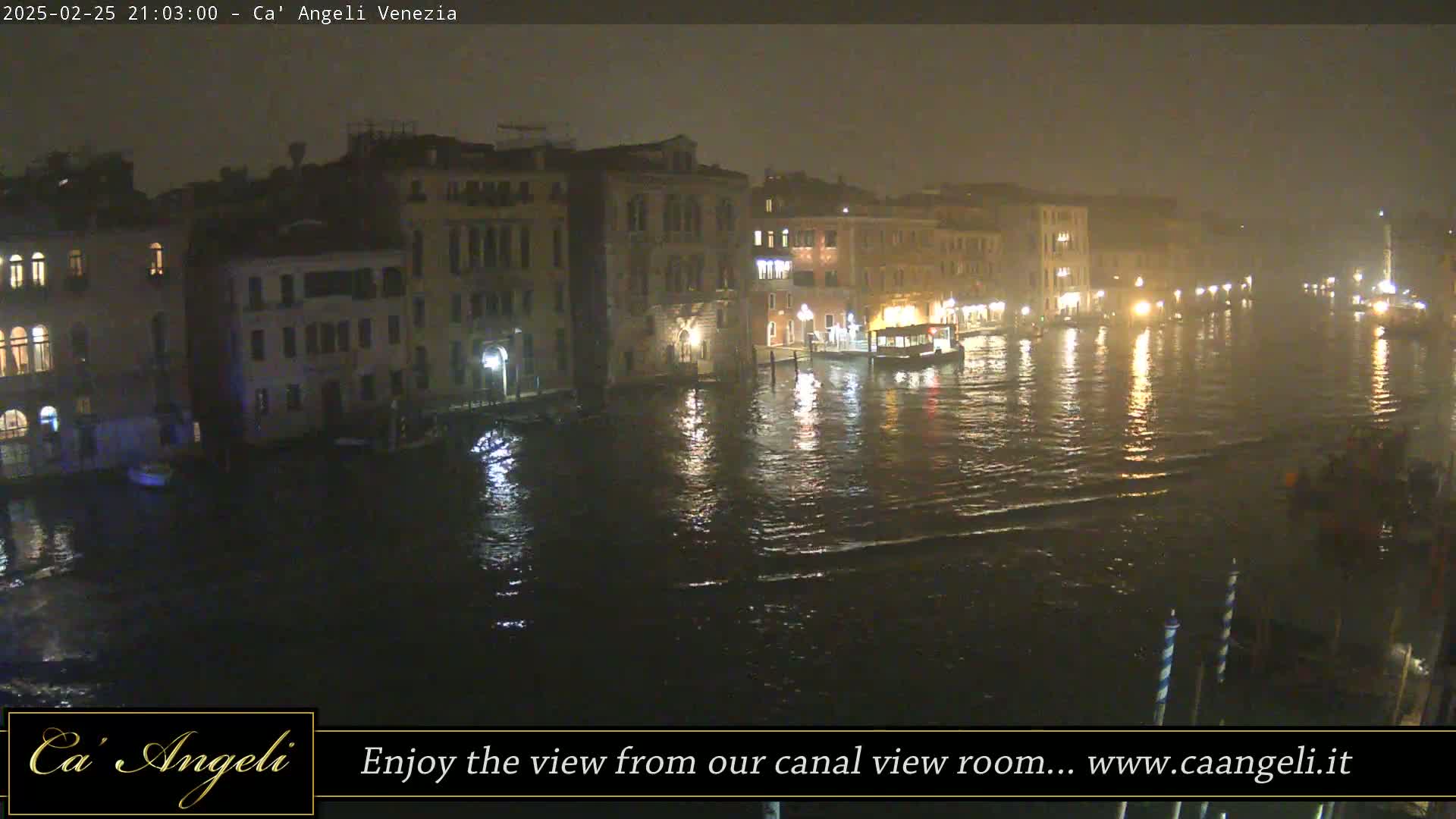 A nighttime view of a canal in Venice, Italy, showing several buildings along the waterway under misty or foggy conditions with some boats on the water.
