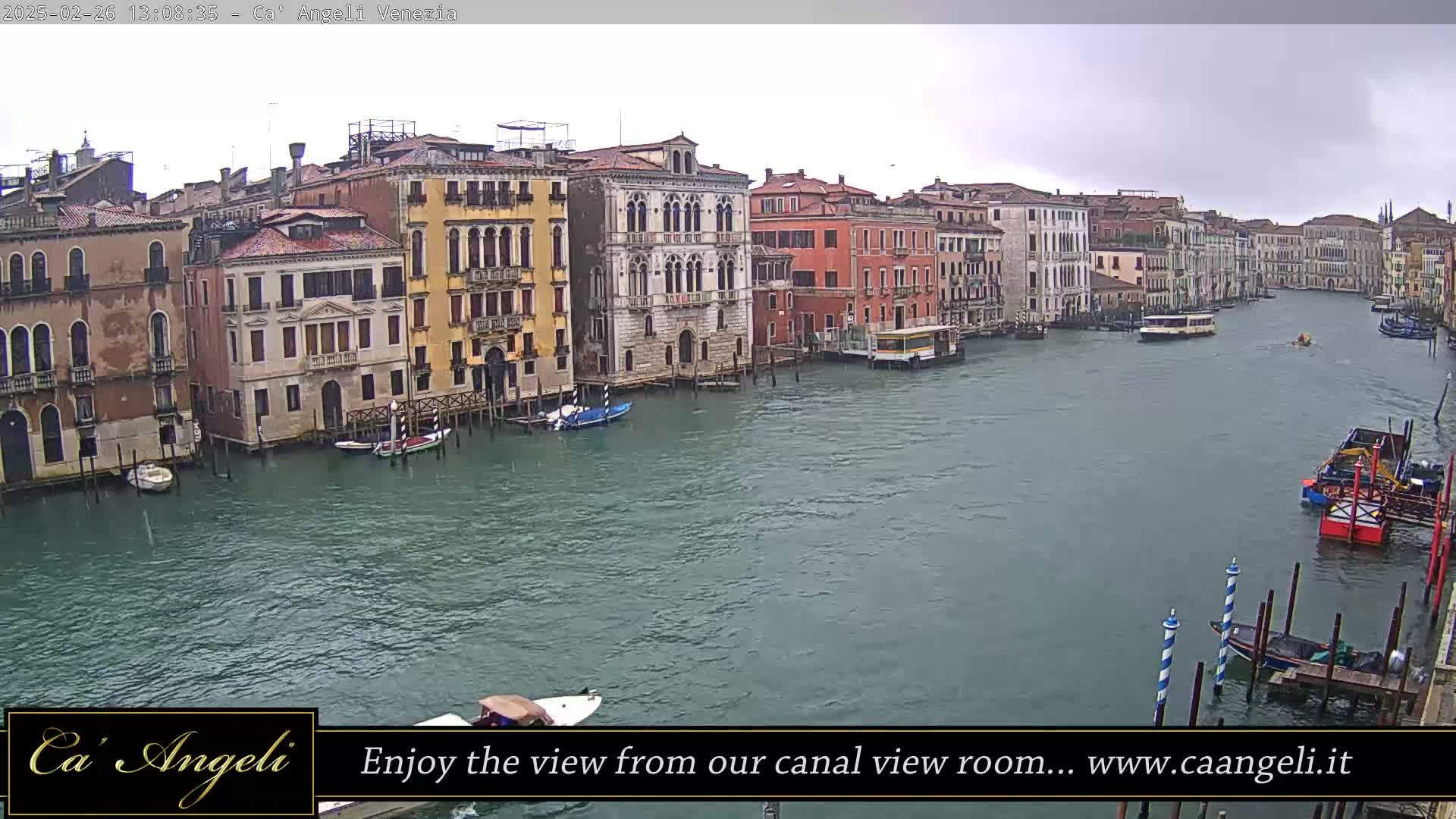 A wide shot of a canal in Venice on an overcast day shows several multi-story buildings lining the waterway, with various boats and watercraft traveling on it.