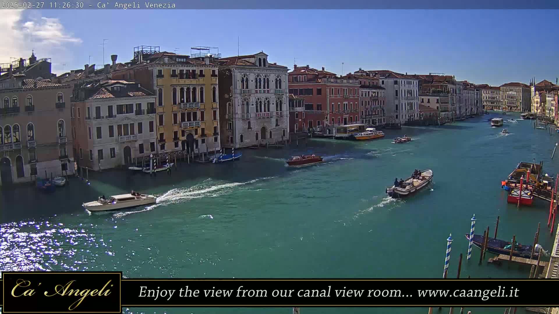 A sunny day in Venice shows several boats traveling on a canal lined with closely-spaced buildings.