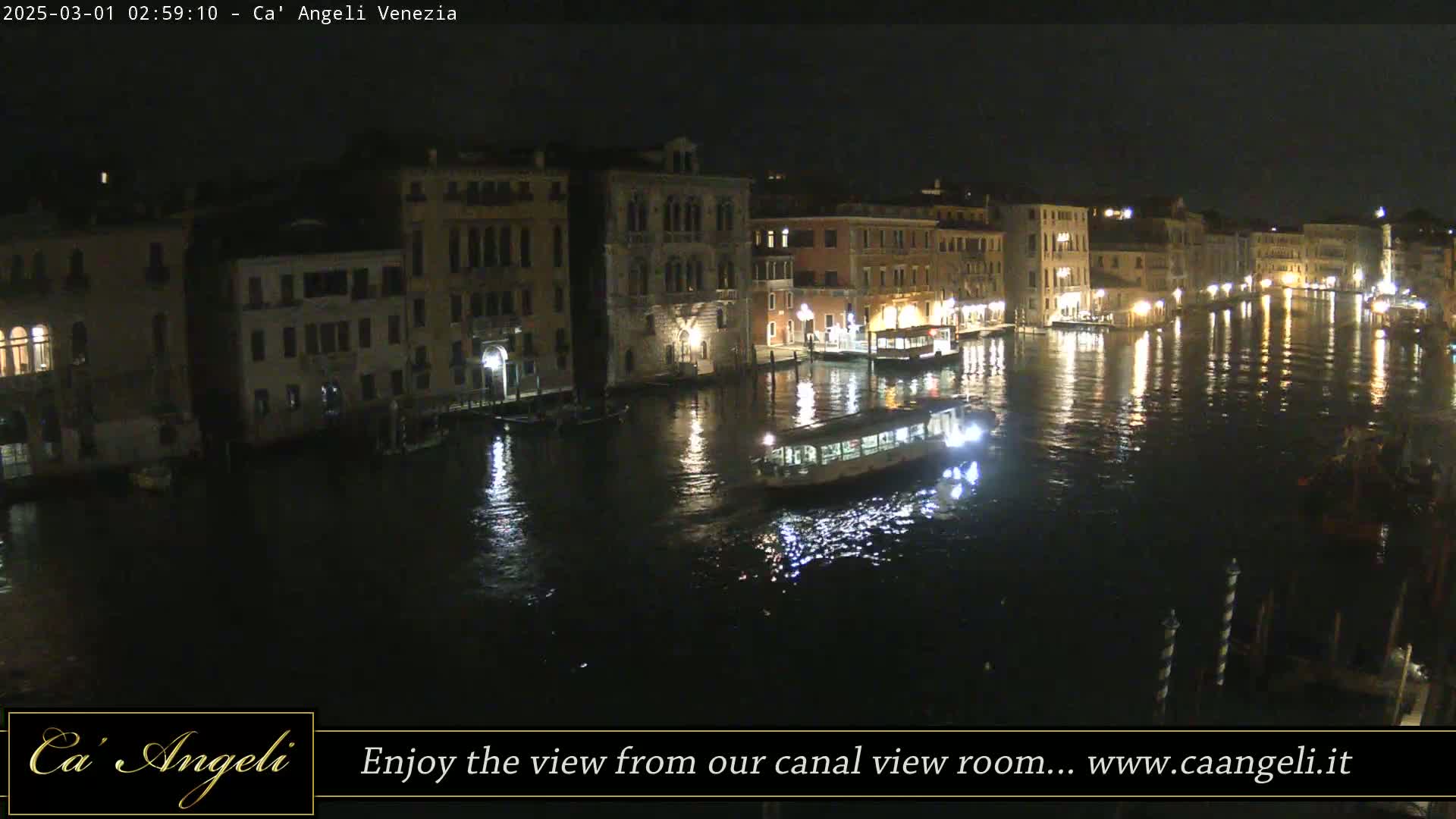 A nighttime view of a canal in Venice, Italy, shows illuminated buildings lining the waterway, with a boat traversing the dark water.