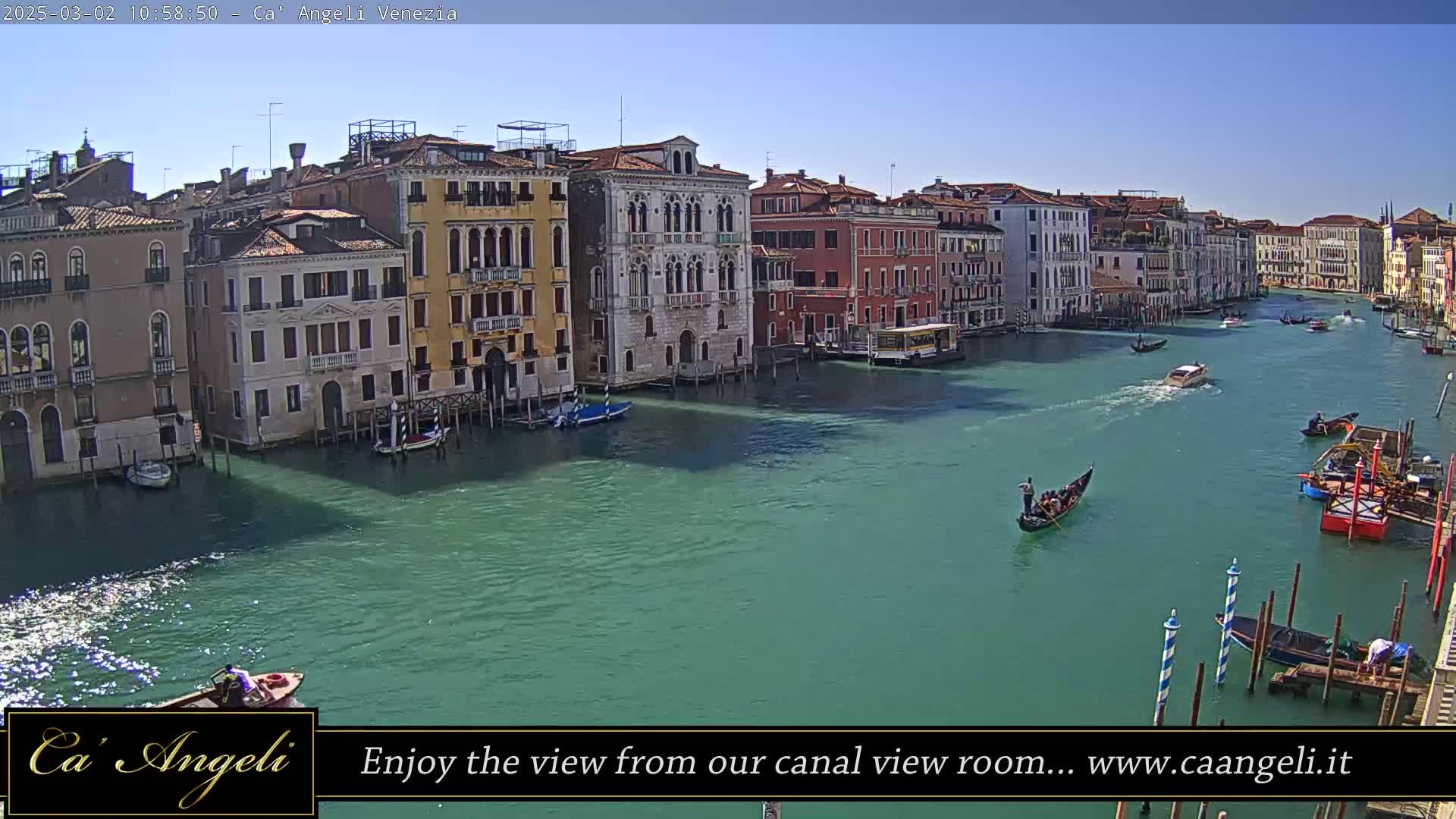 Under a clear blue sky, a wide canal in Venice is filled with gondolas and other small boats passing by colorful buildings along its banks.