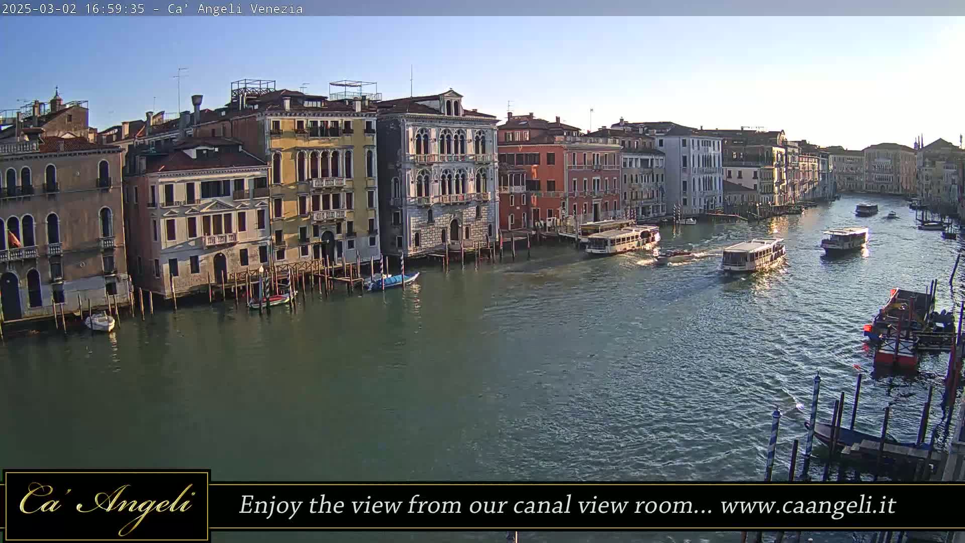 Several boats travel down a canal lined with tall, closely situated buildings under a clear, bright sky.