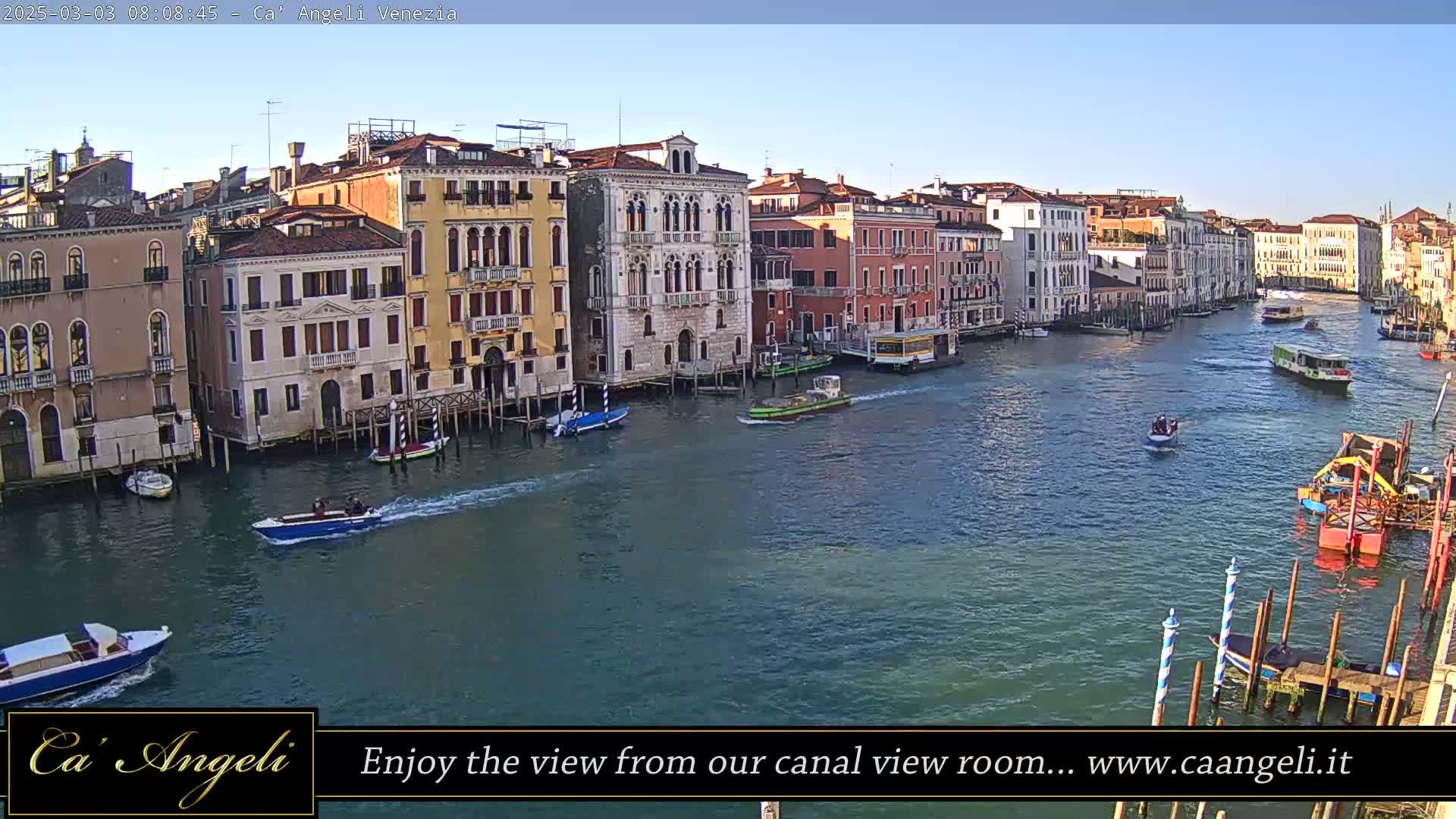 A wide canal in Venice, under a clear sky, is filled with boats passing colorful buildings lining the waterway.