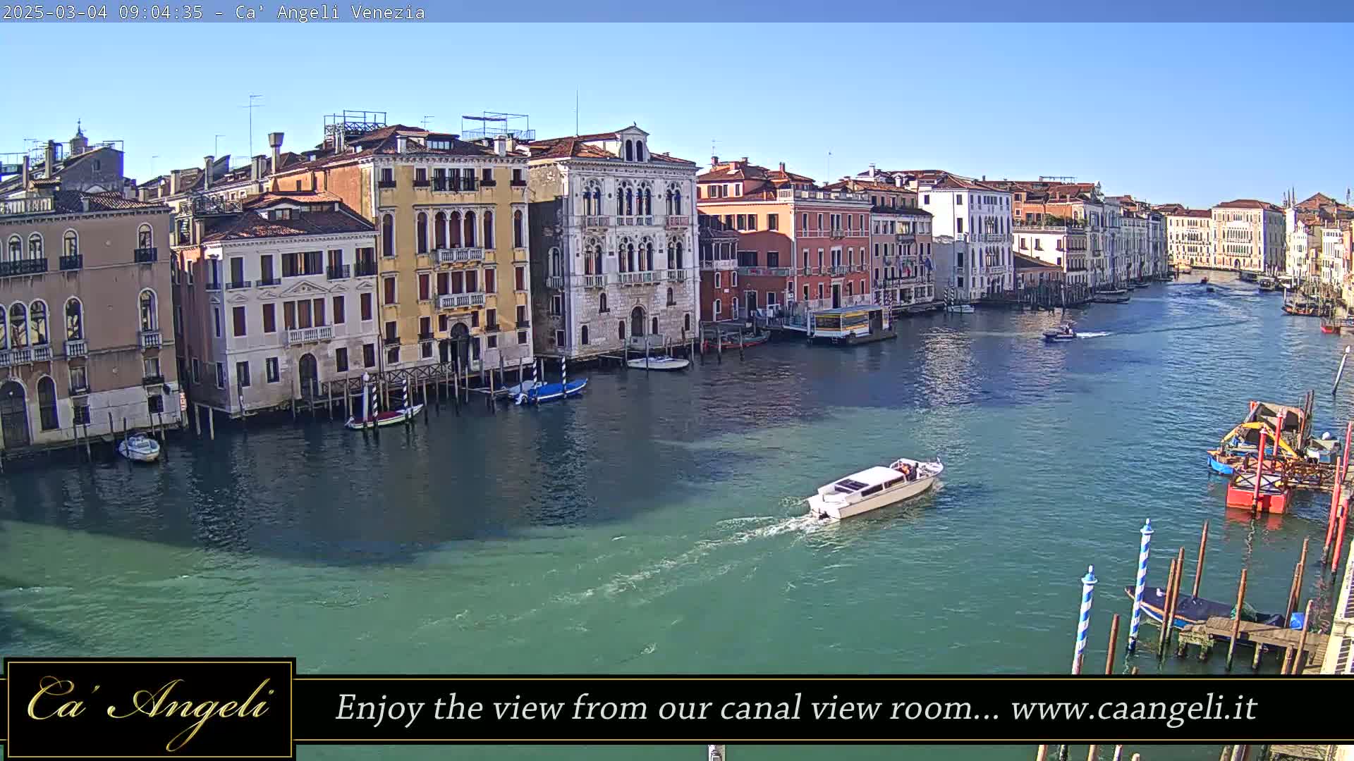 A sunny day in Venice shows a wide canal with colorful buildings lining its banks, several small boats on the water, and a larger motorboat passing by.