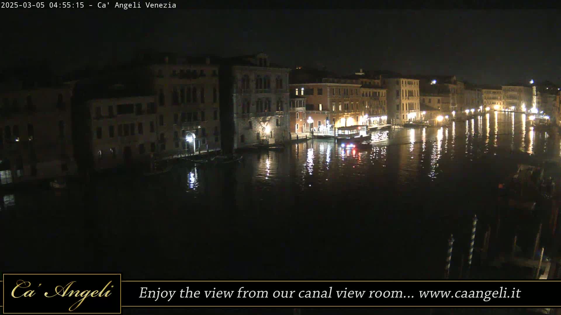 A nighttime view of a canal in Venice, showing illuminated buildings along the waterfront and a few boats on the water.