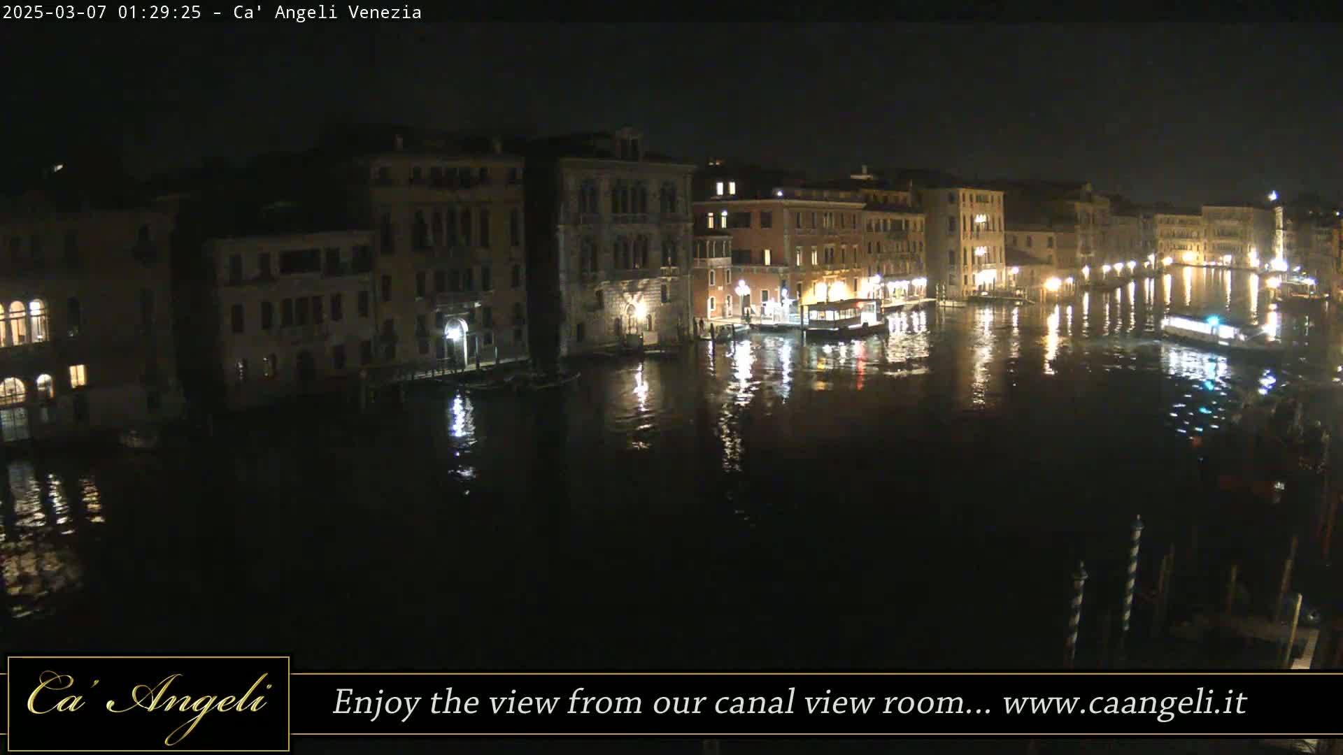 A nighttime view of a canal in Venice, Italy, showing illuminated buildings lining the waterway with boats moored along the shore.