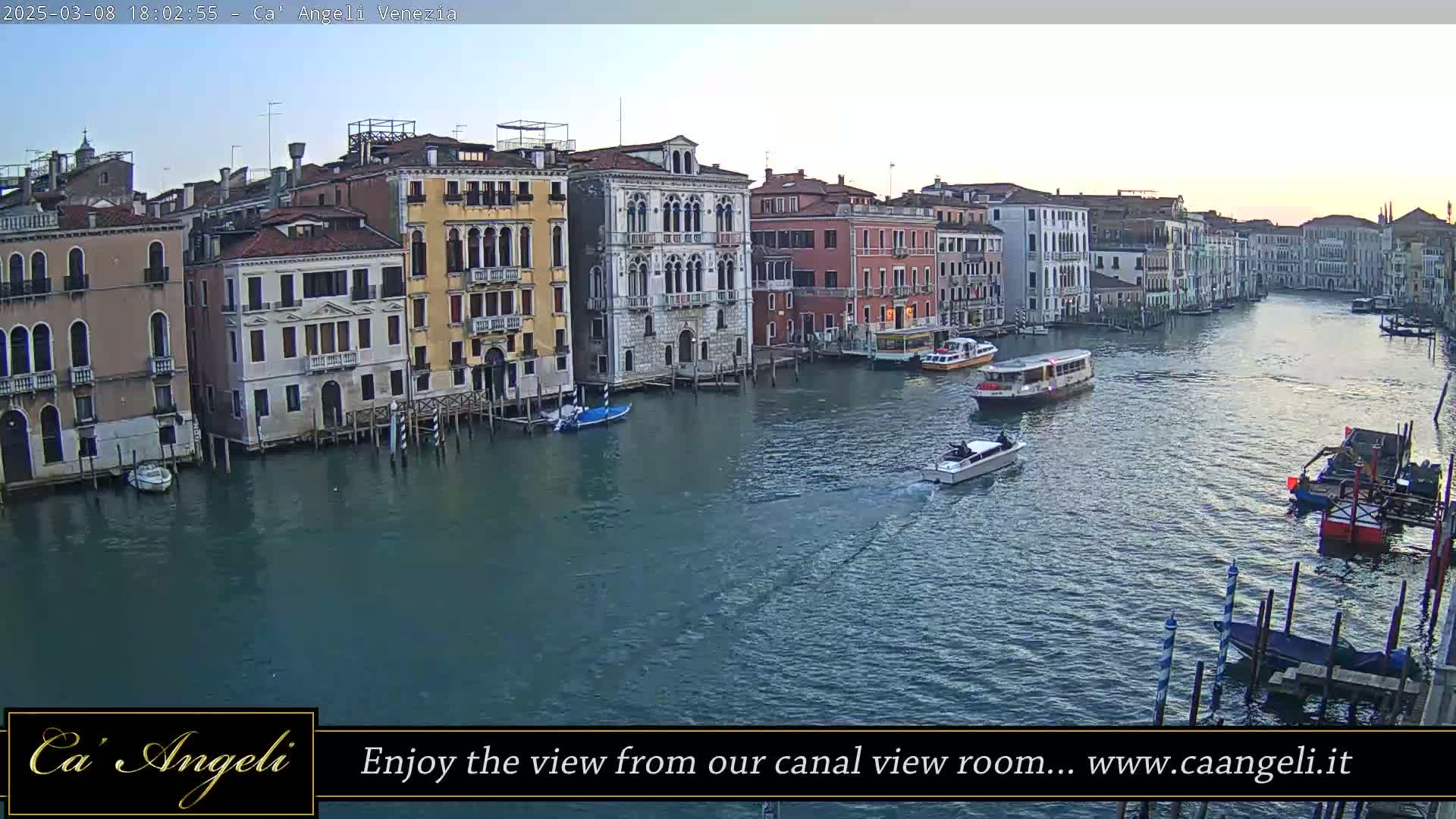 A wide shot of a canal in Venice at dusk, showing colorful buildings lining the waterway with boats passing by.