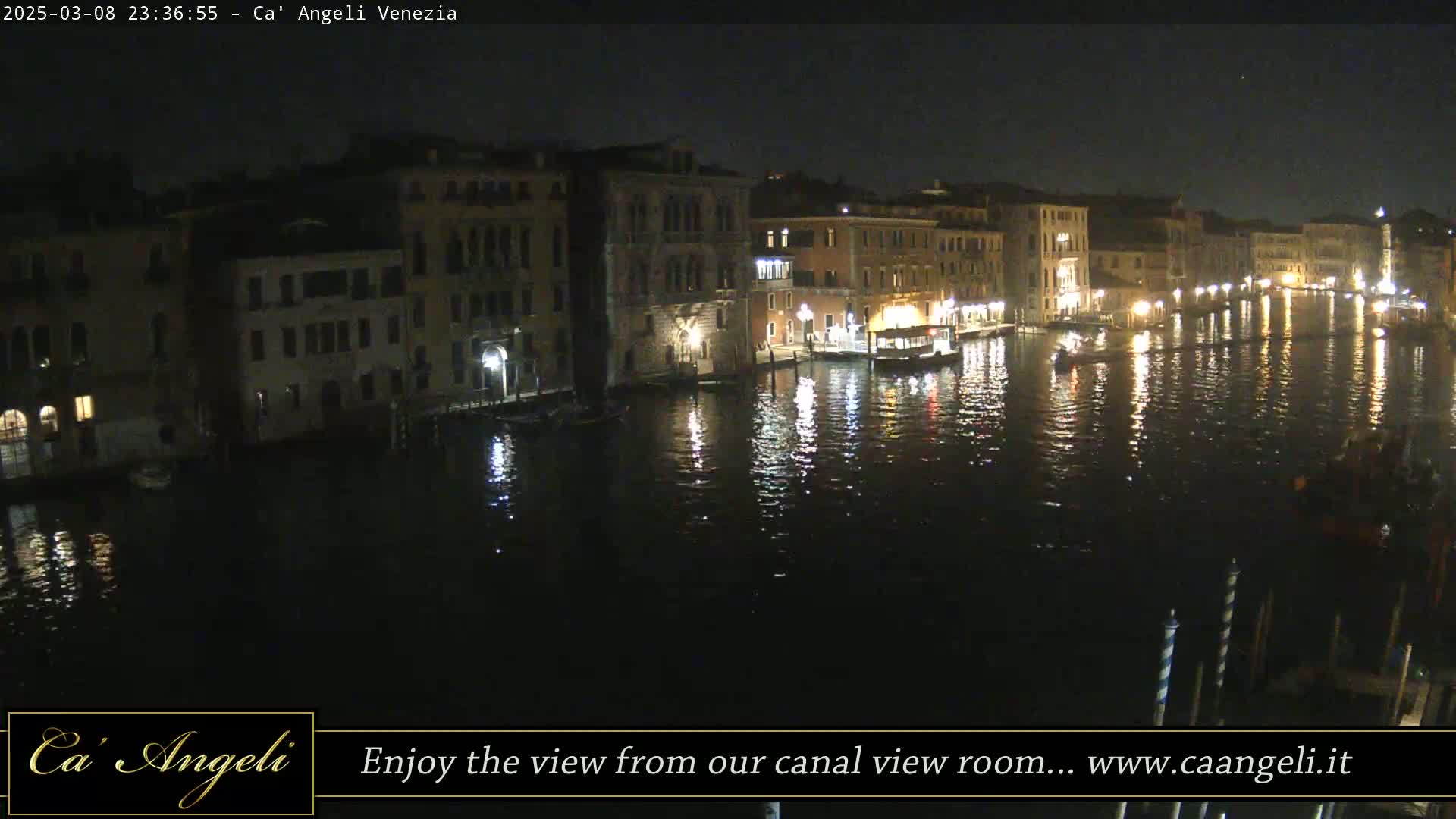A nighttime view of a canal in Venice, showing illuminated buildings lining the waterway, with reflections shimmering on the water's surface under a clear, dark sky.