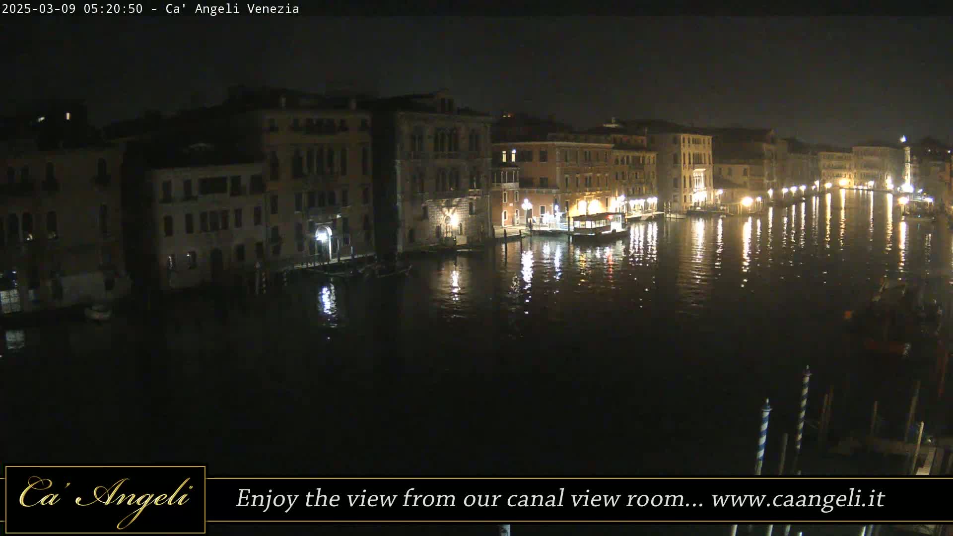 A nighttime view of a canal in Venice, showing illuminated buildings lining the water's edge, with their lights reflected in the calm water.