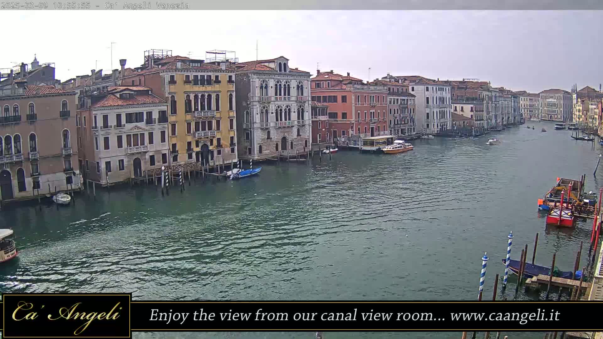 A wide shot of a canal in Venice, Italy on an overcast day, shows several buildings lining the waterway with boats moored along the edges and others traveling down the center.