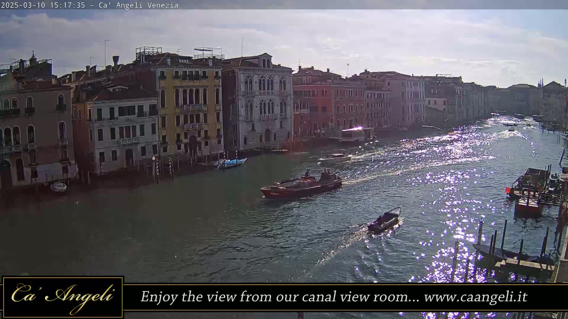 A sunny day in Venice shows a wide canal with several boats, lined with tall buildings.