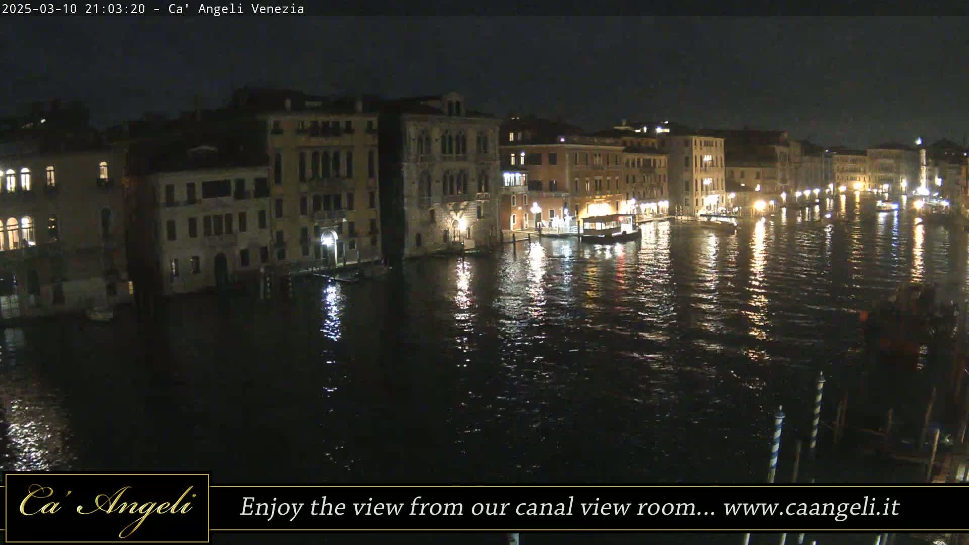 A nighttime view of a canal in Venice, showing illuminated buildings lining the waterway and a few boats on the water.