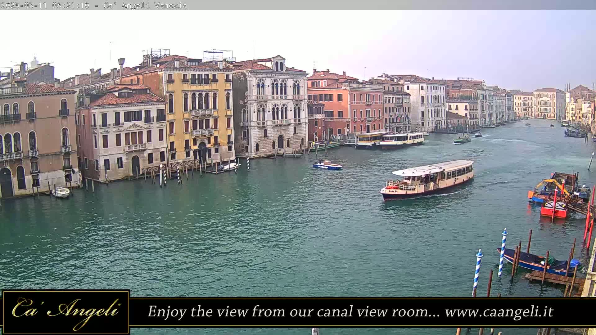 A wide shot of a canal in Venice, Italy, shows several buildings lining the waterway under an overcast sky, with boats and a water taxi moving along the water.