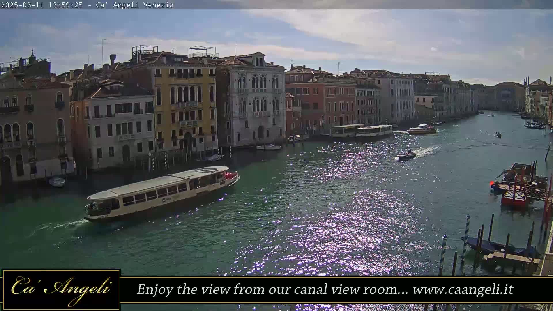 A waterway filled with boats passes by several buildings under a partly sunny sky.