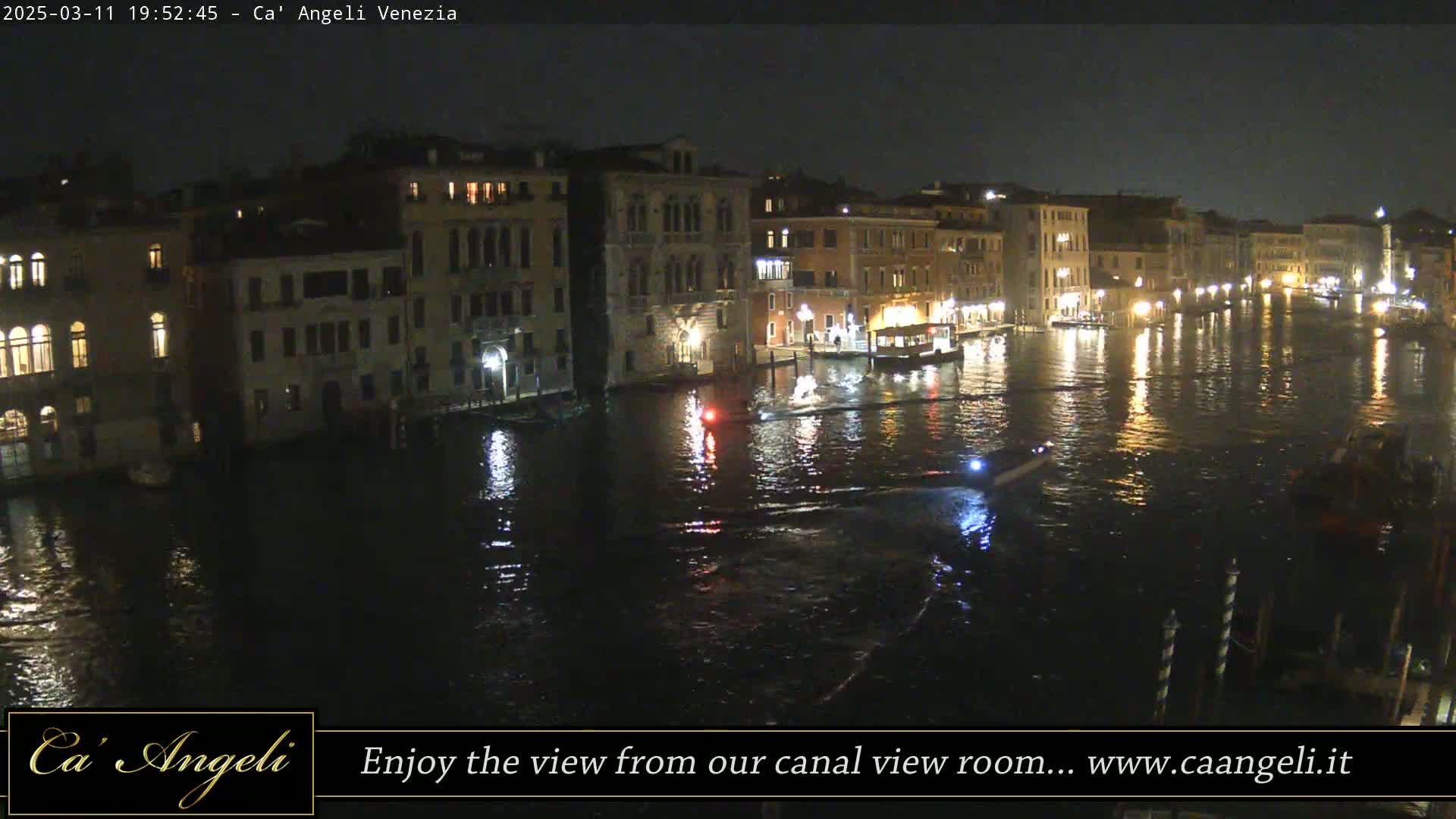 A nighttime view of a canal in Venice, Italy, with illuminated buildings lining the waterway and several small boats on the water.